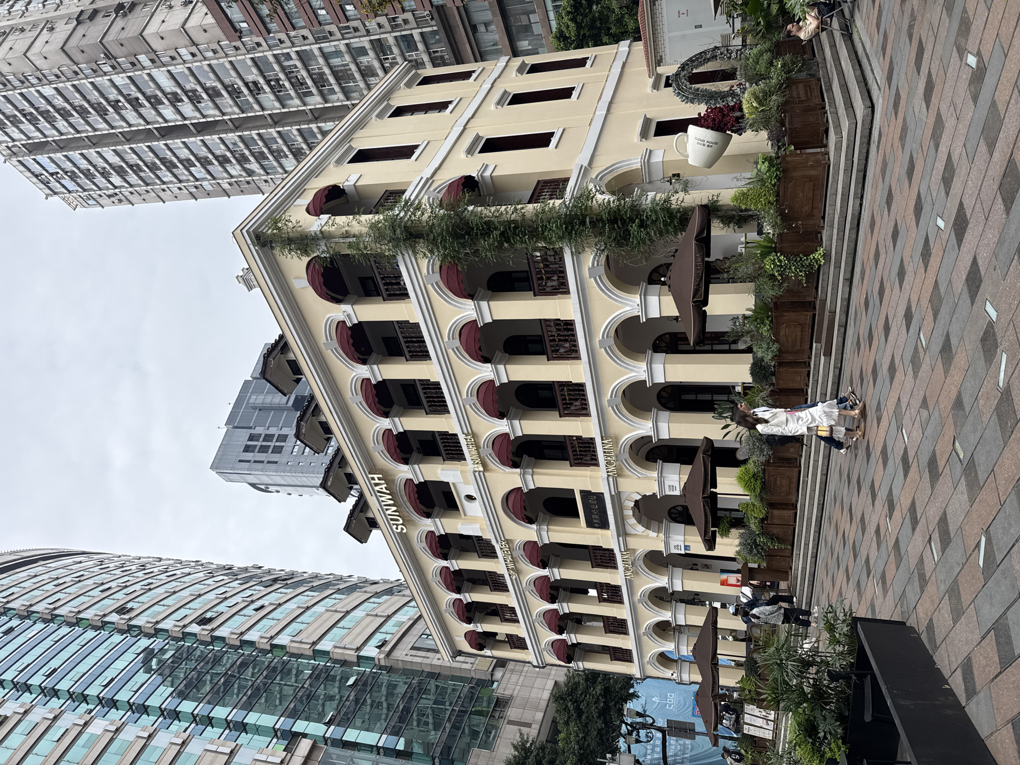 The image shows a European-style building in Yuzhong District, Chongqing, China. The building has arched windows, red awnings, and the words "SUNWAH" and "ANGELINA" on its facade. There are other high-rise buildings in the background. The foreground shows a paved plaza with planters and people walking. A large white mug with text in it is on top of the building.