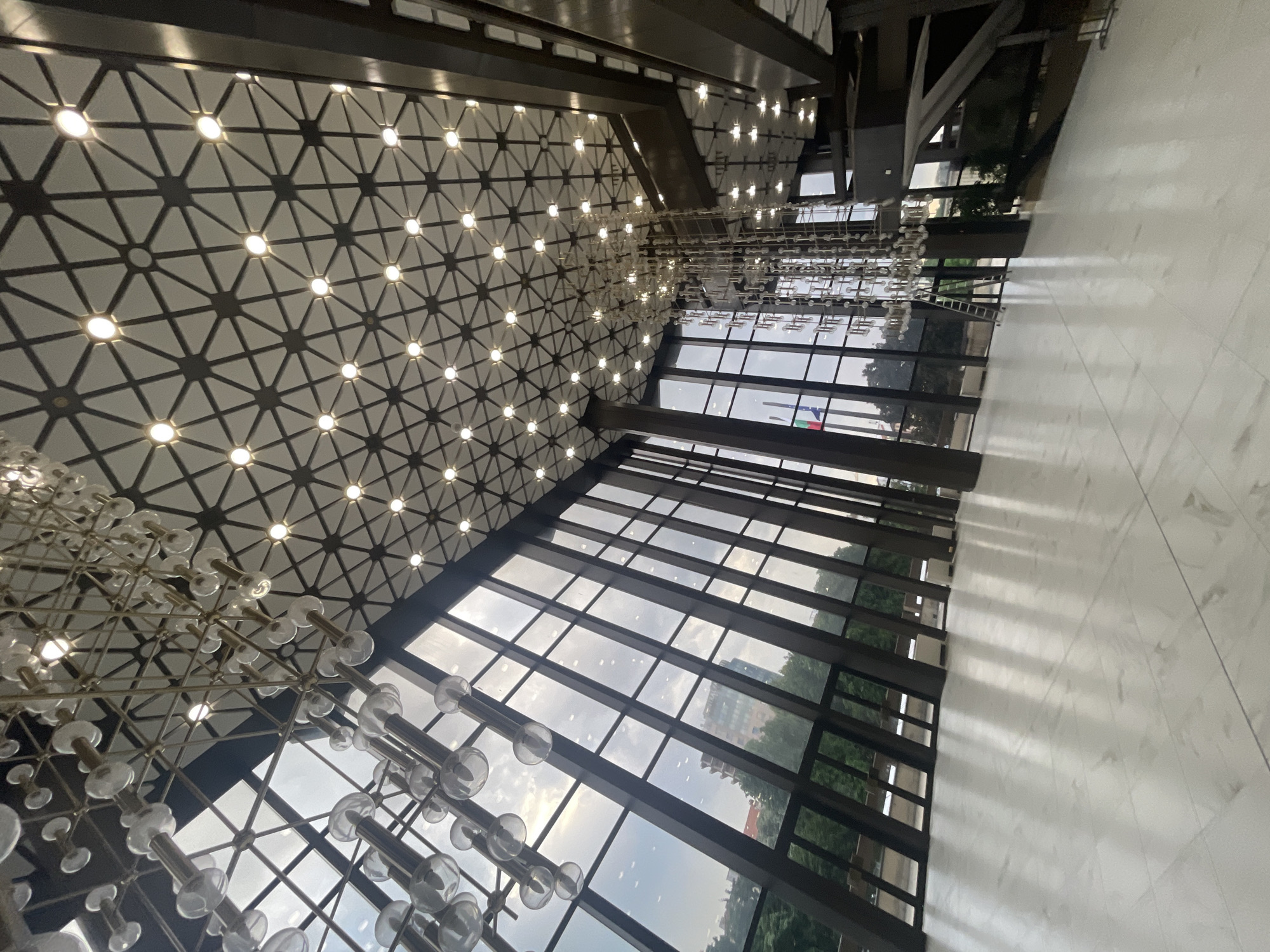 This image depicts the interior of a large, modern hall, identified as the National Palace of Culture (NDK) in Sofia, Bulgaria, evidenced by the distinctive architecture and a visible Bulgarian flag through the window. The perspective is from a low angle, looking upwards and across the expansive space.
The most prominent feature is the ceiling, which is adorned with a complex geometric pattern of dark lines forming interconnected stars, hexagons, and triangles on a lighter, possibly white or off-white, background. Numerous circular spotlights are evenly distributed across this pattern, emitting bright, warm light.
Multiple elaborate chandeliers descend from the ceiling. These chandeliers are composed of numerous individual, clear glass spheres or cylinders, suspended at various lengths by metallic rods, forming a cascading, sculptural arrangement. One particularly long chandelier hangs centrally, extending significantly downwards.
The left side of the hall is dominated by a massive, sloped glass facade, comprising tall, rectangular window panes framed by dark, sturdy vertical and horizontal beams. Through these windows, an exterior view is visible: an overcast sky with visible cloud formations, abundant green foliage from trees, and a distant multi-story building partially visible. A flagpole bearing the Bulgarian flag (vertical white, green, and red stripes) is also seen through the windows on the left.
The floor is composed of large, light-colored, possibly white or light gray, polished tiles with subtle veining patterns. The highly reflective surface of the floor mirrors the ceiling lights, the chandeliers, and the window panes, creating strong linear reflections.
On the right side of the image, dark, angular architectural elements are visible, suggesting multi-level structures or supporting beams. In one of the cropped views, a tall, silver, foldable ladder is positioned near the base of one of the large chandeliers, indicating possible maintenance or installation activity.
The space is devoid of people, presenting a grand, empty interior.