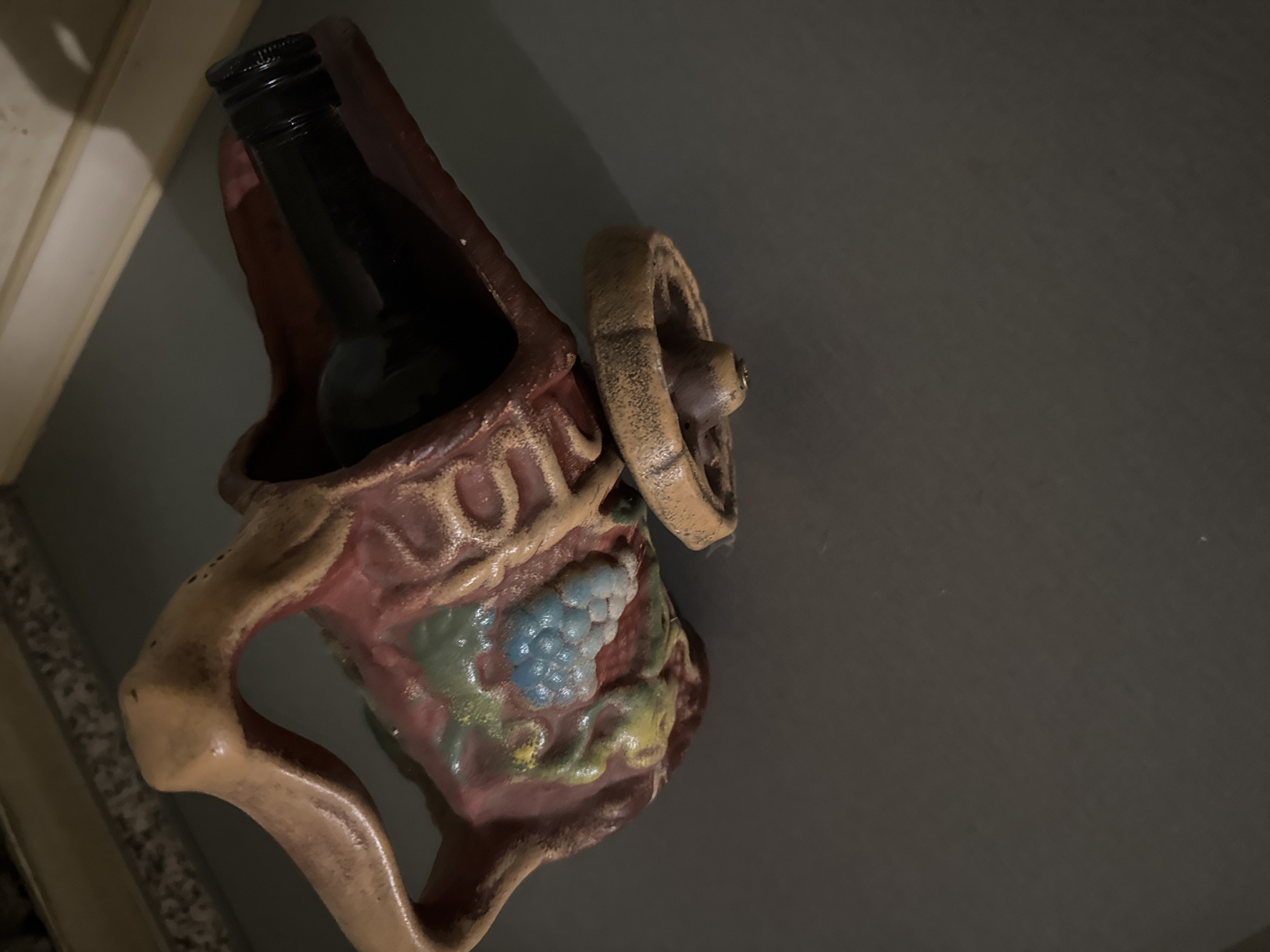 A wine bottle sits inside a decorative holder that resembles a carriage, on a neutral-colored wall. The holder features a wheel on the right side and a handle on the left. A cluster of grapes is painted on the front. The bottle is dark and appears full. No people are visible. There is no explicit information to prove the location is in Yana, Bulgaria.
