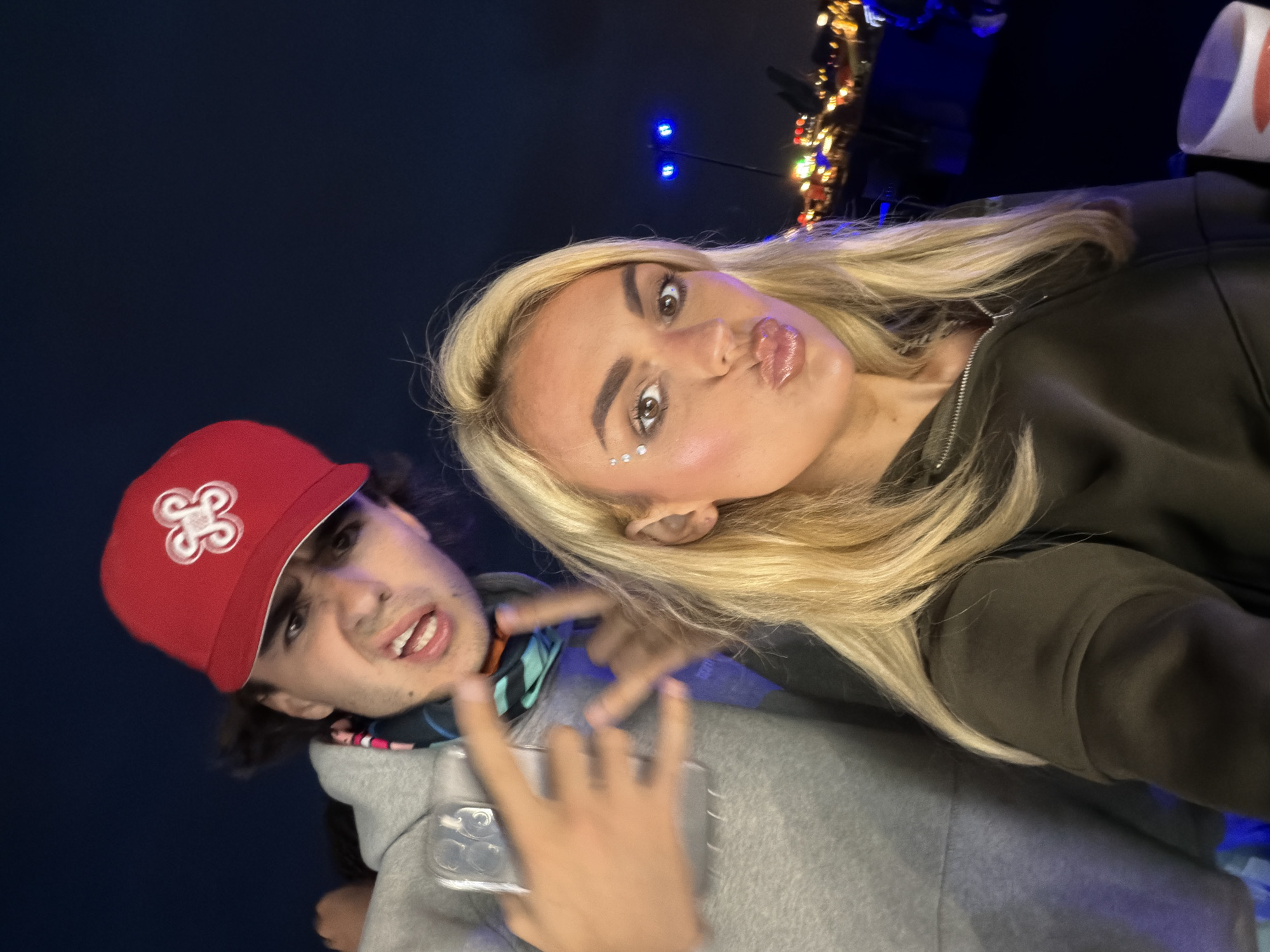 The image is a night-time selfie featuring two young individuals in Sant Adrià de Besòs, Spain. On the left and slightly behind, a male subject with dark hair wears a red baseball cap featuring a white emblem and a grey hooded sweatshirt. A multi-colored neck gaiter with patterns of blue, black, orange, and pink is visible around his neck. His face is contorted in a wide-eyed, open-mouthed expression showing teeth. His right hand, extended towards the camera, displays a "rock on" hand gesture. A clear phone case is visible on his left side. In the foreground to the right, a female subject with long blonde hair wears a dark-colored zip-up jacket. Her makeup includes prominent blush and glossy lips puckered in a "kiss" pose. Several small, reflective embellishments are applied to her face near her left eye. Her eyes are open and directed at the camera. Her right hand, partially visible at the bottom right, holds a white cup with a red design. The background is a dark blue night sky, with blurred, distant city lights and a distinct blue-lit structure on a pole in the middle distance.