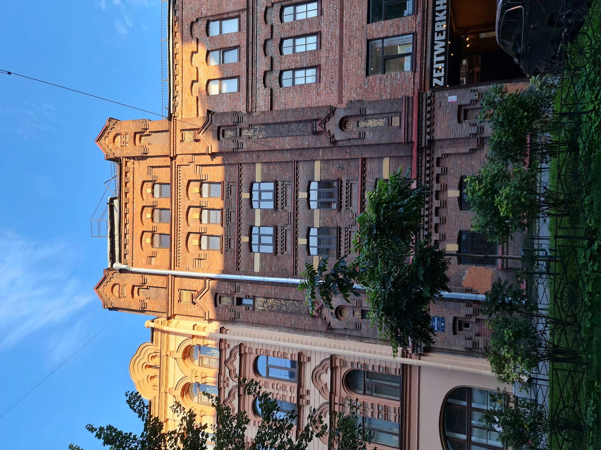 The image depicts the facade of a brick building with ornate architectural details in Saint Petersburg, Russia. The building appears to be in a historic district, with a mixture of architectural styles evident. The building on the left is lighter in color, possibly stucco, with arched windows and decorative elements. The central and right sections are constructed of red brick, featuring rows of windows, brickwork detailing, and a prominent vertical pipe. The upper portion of the central brick building has a tower-like element with arched openings. A sign that reads "ZEITWERKH" is visible on the ground floor of the right section, suggesting a commercial establishment. The foreground shows a green lawn with manicured bushes and a black metal fence. A dark-colored car is parked in front of the entrance to the "ZEITWERKH" establishment. The sky above is blue with scattered white clouds, and the sunlight casts shadows, indicating it is likely late afternoon. The overall scene conveys a sense of urban architectural heritage.