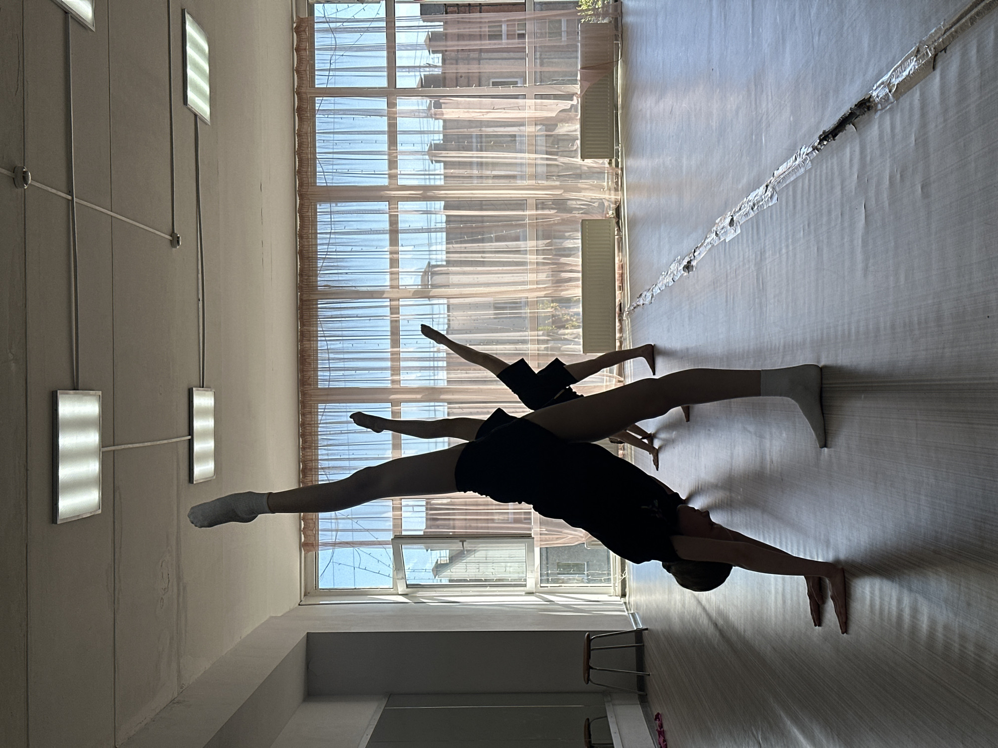 Two dancers are performing in what appears to be a dance studio. The dancers are in the center of the frame, performing a leg split stance. The taller dancer is closer to the camera. They are wearing black shorts, and are holding a split with a wider reach. The other dancer is behind them, also in black shorts. The backdrop is a large window with white, translucent curtains. Outside the window, glimpses of brick buildings suggest an urban environment. Inside, two rows of rectangular lights hang from the ceiling. There are white walls and a reflective light floor. A row of chairs are to the side of the picture. A possible location is Votkinsk, Russia, as identified by the user.