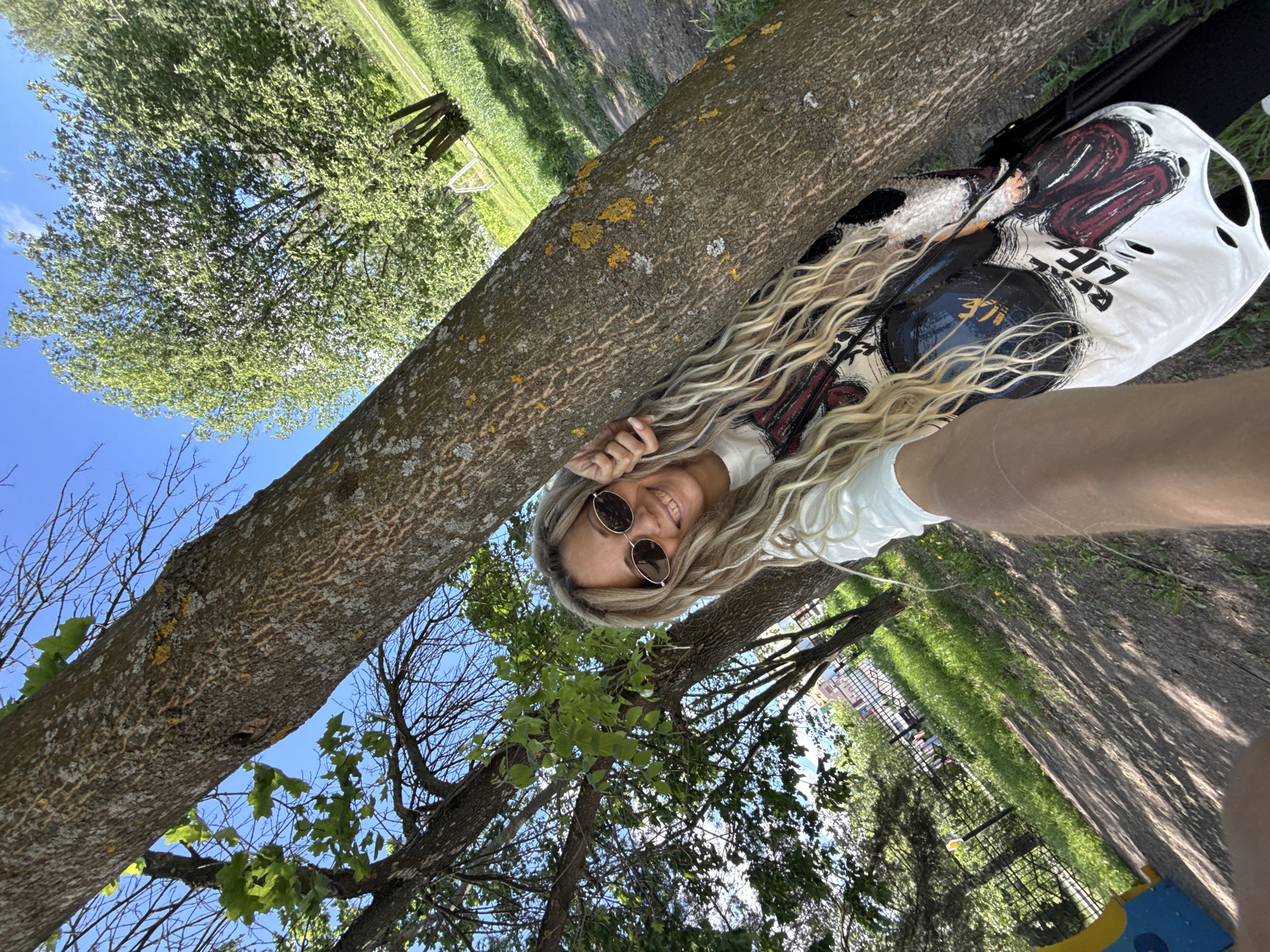 A fair-skinned woman with long blonde hair and sunglasses lies on a tree branch in a park, smiling and looking at the camera. She is wearing a white t-shirt. Her right arm is extended towards the camera. Behind her and to the right, there is a white t-shirt with black graphics hanging on the tree branch. The tree has rough bark and is surrounded by green foliage and other trees, with a blue sky visible through the branches. In the background, there is a grassy area and other park elements visible, possibly including children's playground equipment and buildings. The scene appears to be taken in Rusanovo, Russia, based on the user-provided city name.