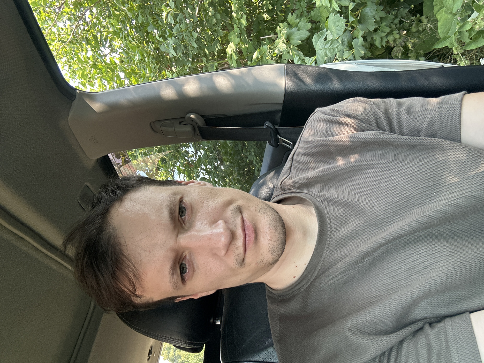 A man with short, dark hair and stubble is seated in the driver's seat of a vehicle. He is wearing a dark grey t-shirt. The interior of the car is visible, including the headrest of the passenger seat and a seatbelt. Outside the car window, lush green foliage suggests a rural or natural setting. The lighting indicates it is daytime, possibly late afternoon, with sunlight casting shadows. The man's expression is neutral. There is no indication of the location being Shatava, Ukraine.