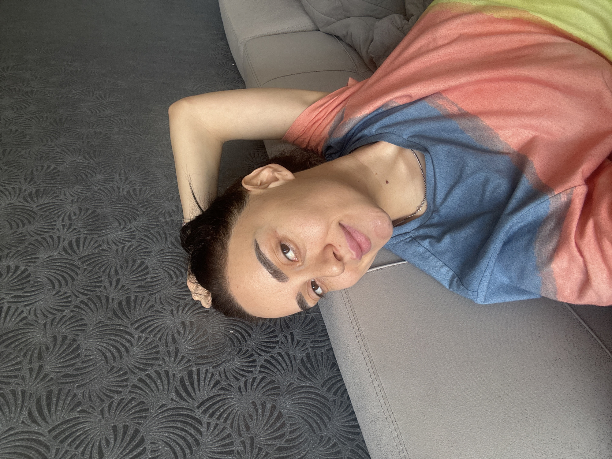 An adult individual is lying on a light gray sofa, looking up at the camera with a playful expression, her tongue slightly extended. She has dark hair, styled away from her face, prominent eyebrows, and brown eyes. She is wearing a multi-colored top that transitions from blue at the bottom, to coral/pink in the middle, and then to a bright yellow/lime green at the top. A silver-colored chain necklace is visible around her neck. Her right arm is bent with her hand seemingly resting behind or above her head, on the sofa. The sofa occupies the right side of the frame, while the left side is covered by a dark gray carpet featuring a detailed, swirling fan-like or abstract floral pattern. The setting appears to be indoors, likely a relaxed home environment in Tbilisi, Georgia. The lighting is soft and even, suggesting an indoor setting with either artificial light or diffuse natural light. There is no visible text in the image.
