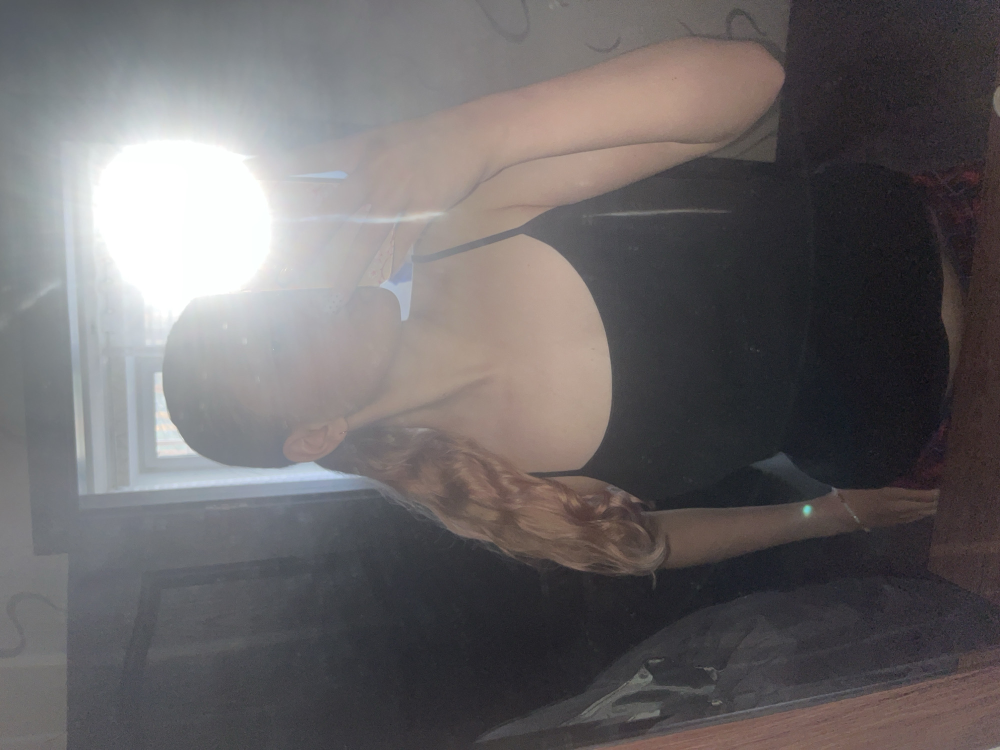 The image captures an indoor scene, likely a room, with a person as the primary subject. The person appears to be a female with fair skin and long, wavy or curly reddish-blonde hair cascading over her shoulder. She is wearing a dark, possibly black, top with thin straps. A thin bracelet is visible on her right wrist.
A strong, bright light source, likely from a window on the left side of the frame, dominates the scene, creating a large, intense glare that partially obscures the upper part of the person's head and the immediate background. This backlighting leaves much of the foreground and the room in deep shadow. The person is positioned in a way that suggests a pose for a photograph, with her body angled and one arm raised, possibly holding a device or simply part of the pose.
Notable details include the extreme brightness from the window, indicating it is likely daytime. The strong glare makes it difficult to discern specific features of the room or the person's expression. No discernible text is visible in the image. While the location context is provided as Perm, Russia, there are no specific visual cues within the image itself to confirm this.