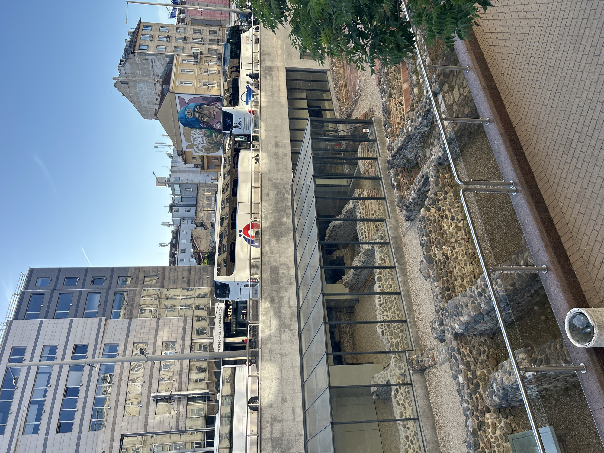 The image depicts a section of the Ancient Serdica Archaeological Complex in Sofia, Bulgaria. Preserved Roman ruins are visible behind a glass enclosure and along the street level, made of stone and concrete, in an open-air museum. A modern building with reflective glass windows is located next to the ruins and buses pass by. The sidewalk is made of pavers and lined with a metal handrail.