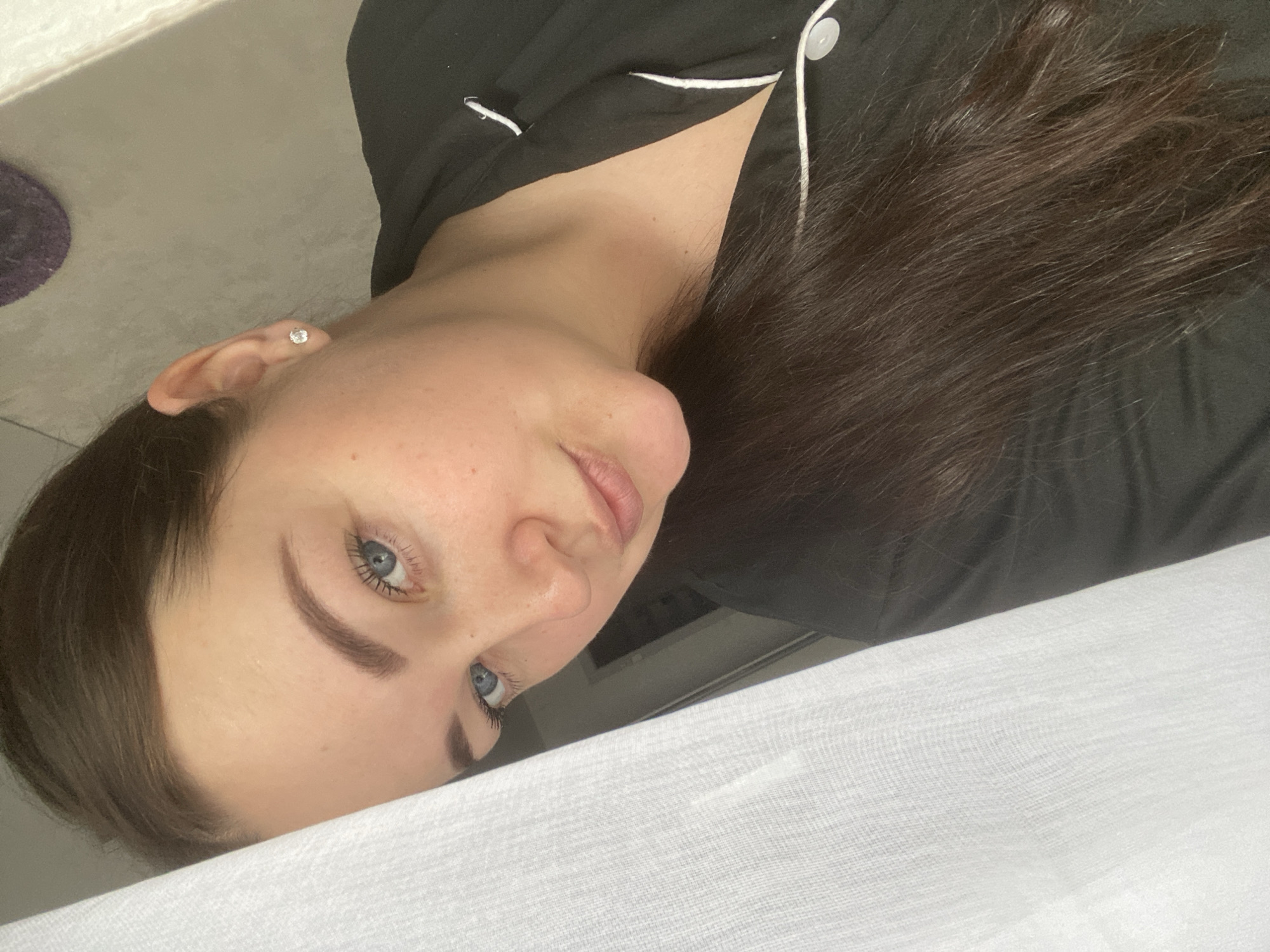 The image is a close-up of a woman lying on a white surface, likely a bed. She has blue eyes, brown hair, and is wearing a dark-colored top with white piping, possibly pajamas. The background is out of focus, but glimpses of a wall and what appears to be a small object on the floor are visible. There is no indication of the city of Sochi, Russia.