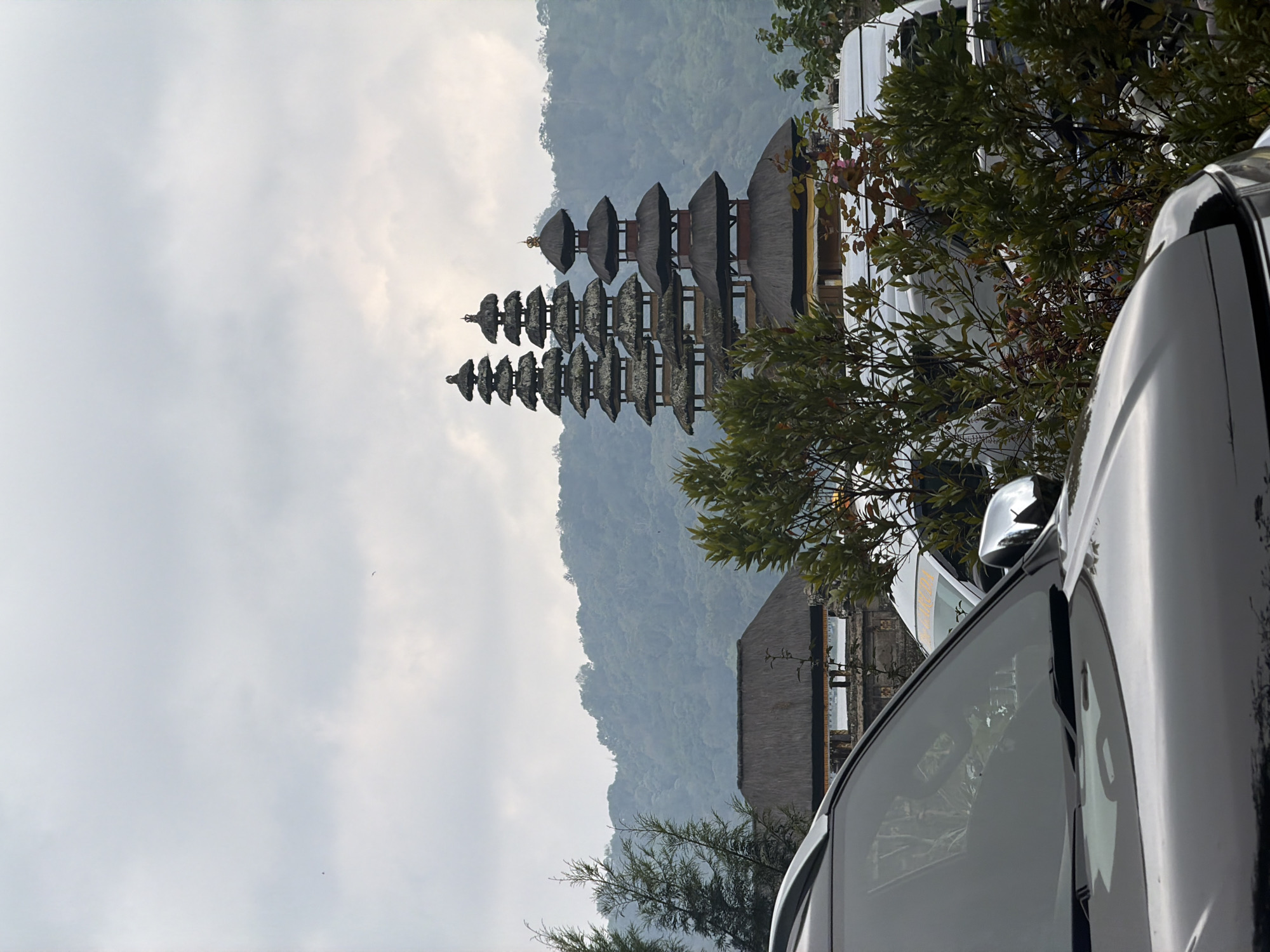 In Banjar, Indonesia, on a cloudy day, a group of tiered temples with thatched roofs stands against a misty green mountain backdrop. In the foreground, the hood and windshield of a black car obscure a portion of the scene, reflecting the overcast sky. To the left of the temples, a structure with a thatched roof is visible, and to the right, lush green foliage partially conceals other vehicles, including a white van with orange lettering. The overall atmosphere is serene and somewhat diffused due to the weather.