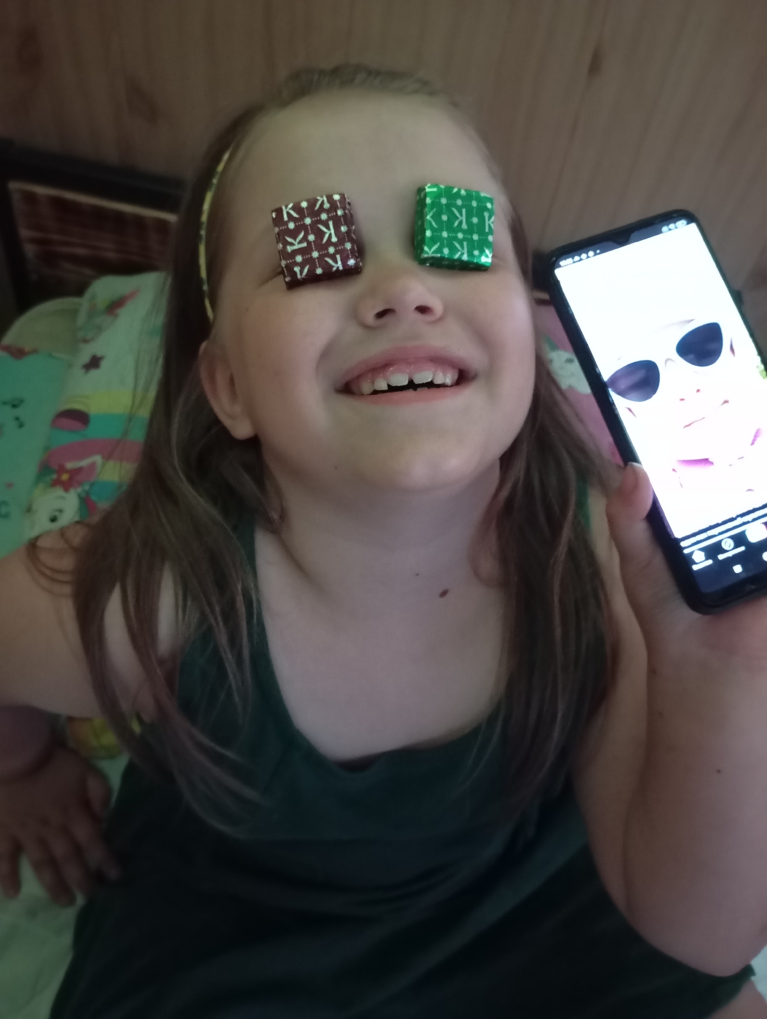 A young girl with brown hair is smiling and looking up. She has two small, square candies on her eyes, one wrapped in green foil and one in brown foil. She is holding a smartphone in her right hand, the screen displaying a cartoon image of a person wearing sunglasses. She is wearing a dark green sleeveless top and a yellow headband. She appears to be indoors, seated on a bed with a patterned blanket. The location is Unknown, Unknown.