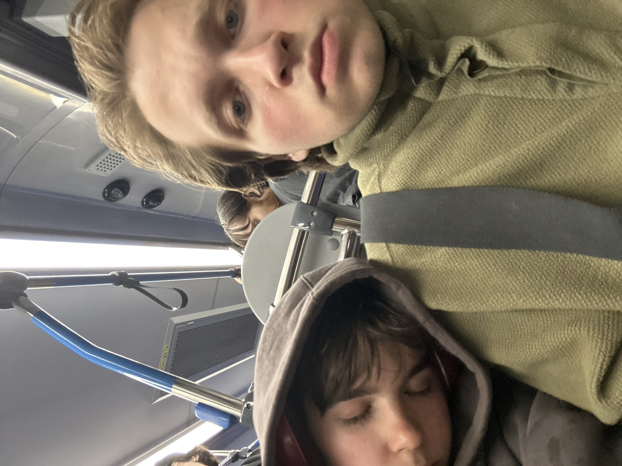 A close-up, selfie-style image shows two young individuals on public transportation. The person in the foreground, facing the camera, has light brown hair and appears to be looking directly at the lens with a neutral expression. They are wearing a textured, olive-green garment. To their left, another individual is asleep, resting their head on the first person's shoulder. This sleeping individual is wearing a dark hooded garment. The background suggests the interior of a bus or train, with metallic handrails, a blue pole, and part of a ventilation grate visible. The lighting is bright, likely from overhead lights. The location is identified as Unknown, Unknown.