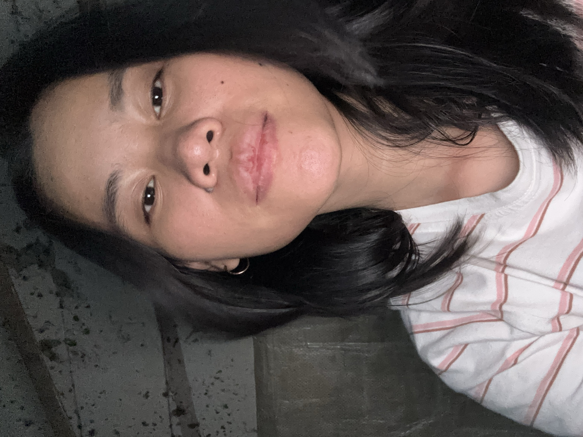 A medium shot shows a young Asian woman from the chest up, looking directly at the camera. She has long, dark hair that falls around her shoulders, framing her face. Her eyebrows are dark and arched, and her eyes are dark brown. She is wearing a white t-shirt with thin pink and brown stripes. The background is dark and appears to be a textured, possibly concrete wall with some debris, suggesting an outdoor or industrial setting. The lighting is dim, casting some shadows on her face and hair. The overall impression is a casual selfie taken in a dimly lit environment.