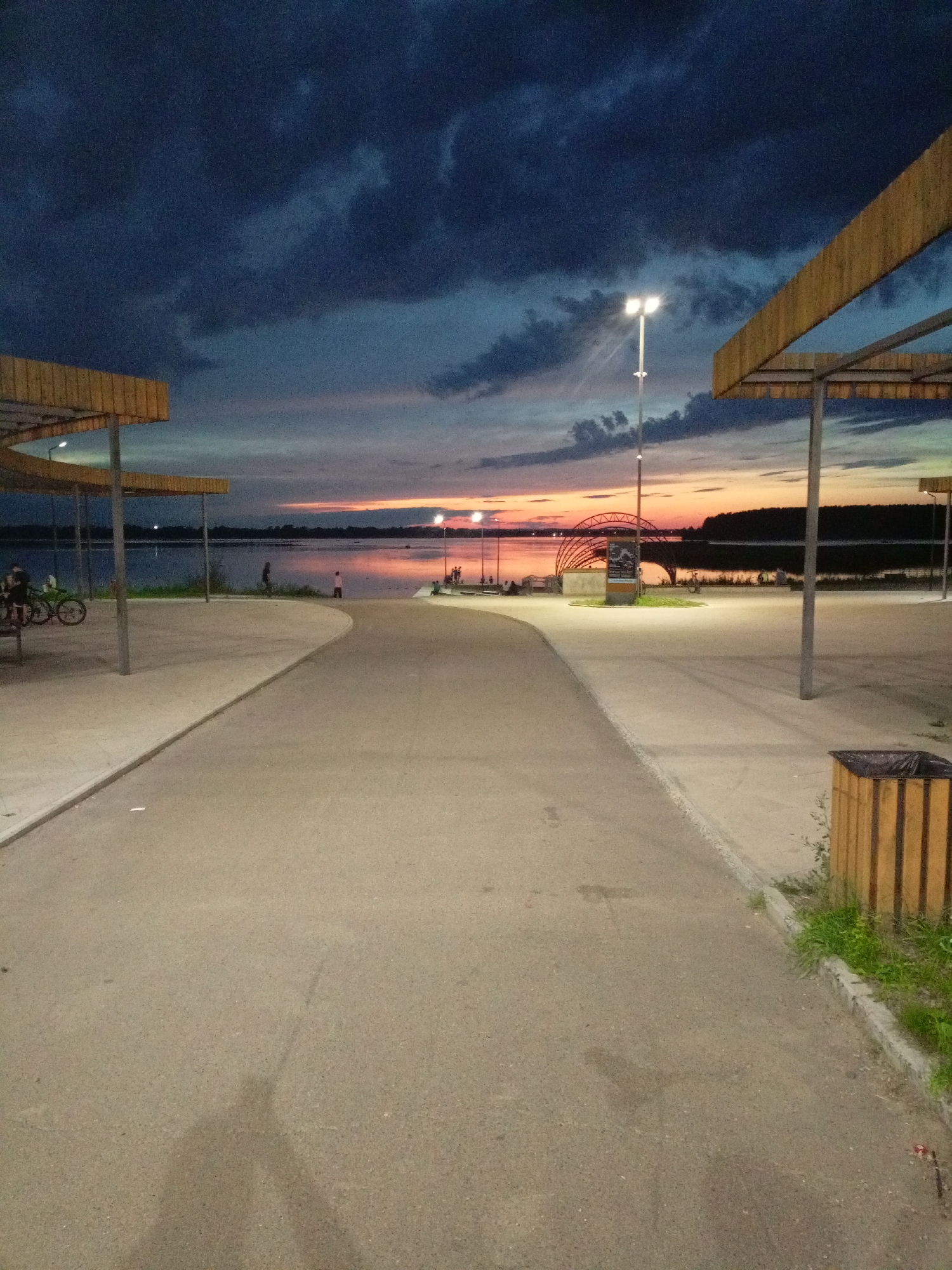 The image depicts a twilight scene at a waterfront promenade, likely in Zheleznogorsk, Russia, given the context. A paved pathway curves towards a body of water, where the sky is a dramatic blend of dark, heavy clouds above and a vibrant sunset with hues of orange and pink closer to the horizon. Several sheltered seating areas with wooden beams and metal supports are visible on either side of the pathway.
To the left, a few bicycles are parked near one of the shelters, and several indistinct figures are present. Further along the waterfront, people can be seen sitting and standing. In the distance, across the water, a treeline forms a dark silhouette against the colorful sky. An architectural feature, possibly a bridge or archway, is visible in the middle distance. Two lampposts are illuminated, casting a soft light on the pathway. A wooden trash receptacle with a dark liner is positioned near the right edge of the frame. The overall impression is of a public space winding down for the evening.