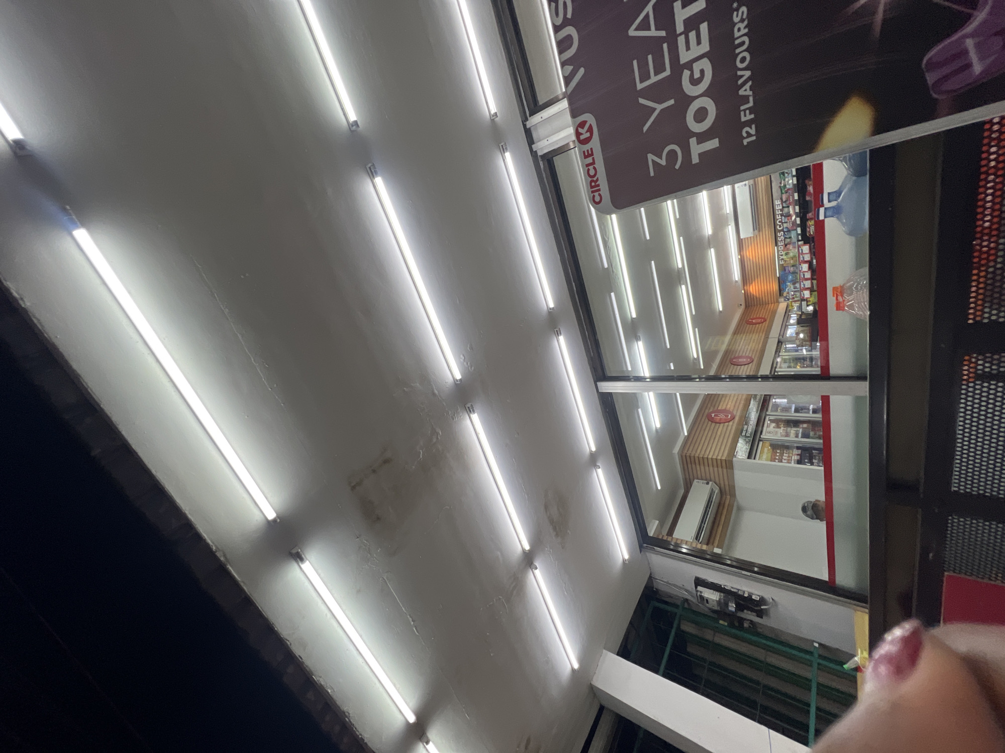 The image depicts the exterior of a Circle K convenience store at night. The facade is illuminated by a series of fluorescent lights lining the ceiling. Through the large glass windows, the interior of the store is visible, showing aisles stocked with products and refrigeration units. A sign on the right side of the image reads "CIRCLE K" and displays promotional text, part of which includes "3 YEARS TOGETHER" and "12 FLAVOURS". In the foreground, a person's finger with pink nail polish is partially visible, obscuring a portion of the view. The environment appears to be an urban setting, with the dark night sky above. The store is located in the city of Denpasar, Indonesia.