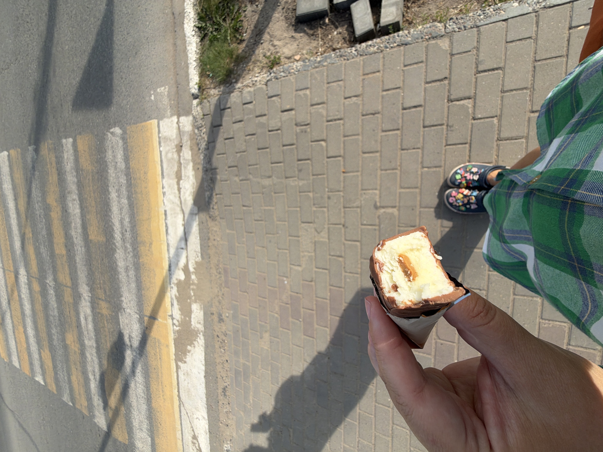 A person is holding a bitten ice cream bar in their right hand. The ice cream has a chocolate coating and a vanilla and caramel filling. The person is wearing a green and white plaid skirt and dark blue slip-on shoes with colorful floral patterns. They are standing on a brick sidewalk next to a crosswalk. The crosswalk has yellow and white stripes. In the background, there is a patch of grass and some construction materials. The scene appears to be in the city of Chaikovsky, Russia, as indicated by the context provided.
