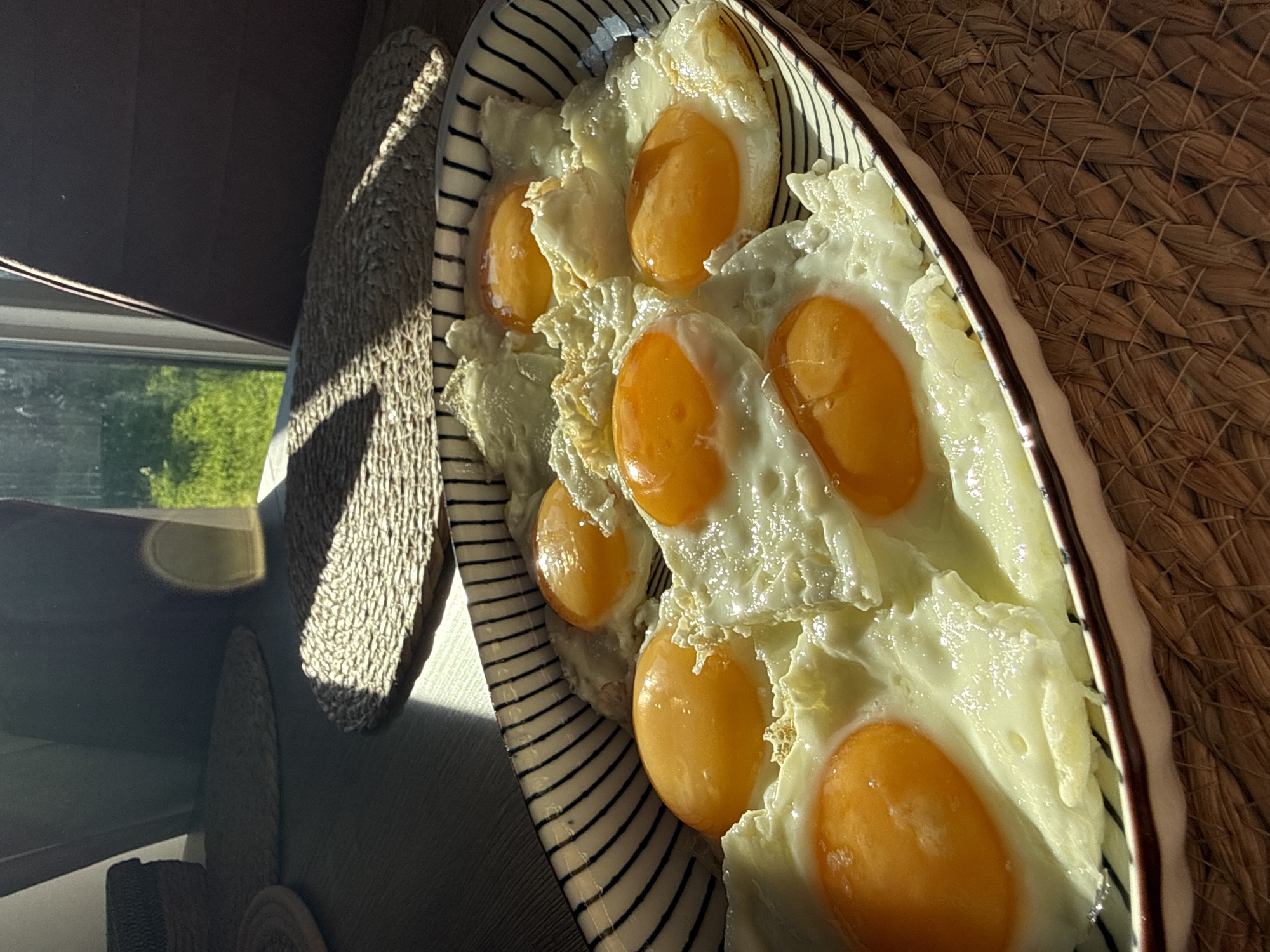 A close-up, eye-level shot showcases a white oval platter filled with seven sunny-side-up fried eggs. The eggs are arranged closely together, with some overlapping. Each egg features a bright orange yolk surrounded by cooked egg white. The platter has a black and white striped rim. The platter rests on a woven straw placemat, which is placed on a dark wooden surface. Sunlight streams in from the right, casting distinct shadows from woven placemats and other objects onto the wooden surface. In the background, a dark object, possibly a chair, is visible, and beyond that, a sunlit green outdoor area suggests a window or doorway. The scene is indoors, and the lighting indicates a bright, sunny day. There are no people or animals present. The location is not specified in the image, and therefore cannot be determined.