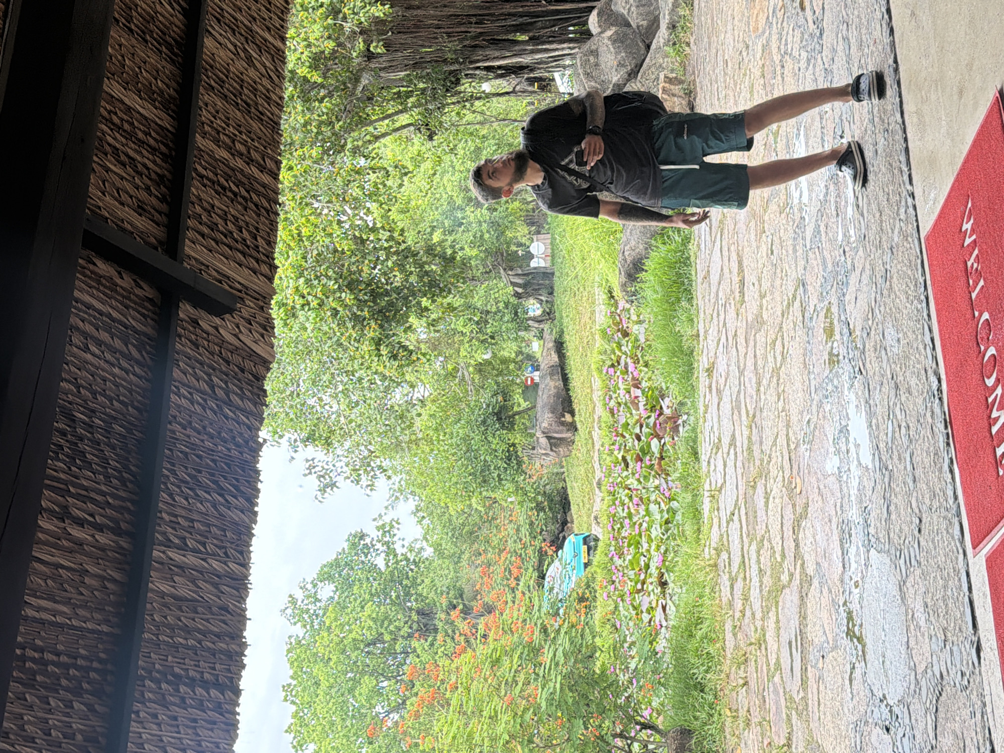 A man with a beard and tattoos stands on a stone patio under a thatched roof. He is looking up and to the right, holding a mobile phone in his right hand. He wears a dark t-shirt and dark green shorts. A black backpack is slung over his left shoulder. The patio is wet, with several puddles reflecting the overcast sky. In the background, a pond filled with pink water lilies is visible, bordered by lush green grass and trees. A turquoise electric scooter is parked near the pond, and orange flowers bloom on a nearby bush. Large rocks are scattered throughout the landscape. A red "Welcome" mat lies on the ground in the foreground. The overall impression is an outdoor area, possibly a botanical garden or a resort in Nha Trang, Vietnam, with a naturalistic and tranquil atmosphere.