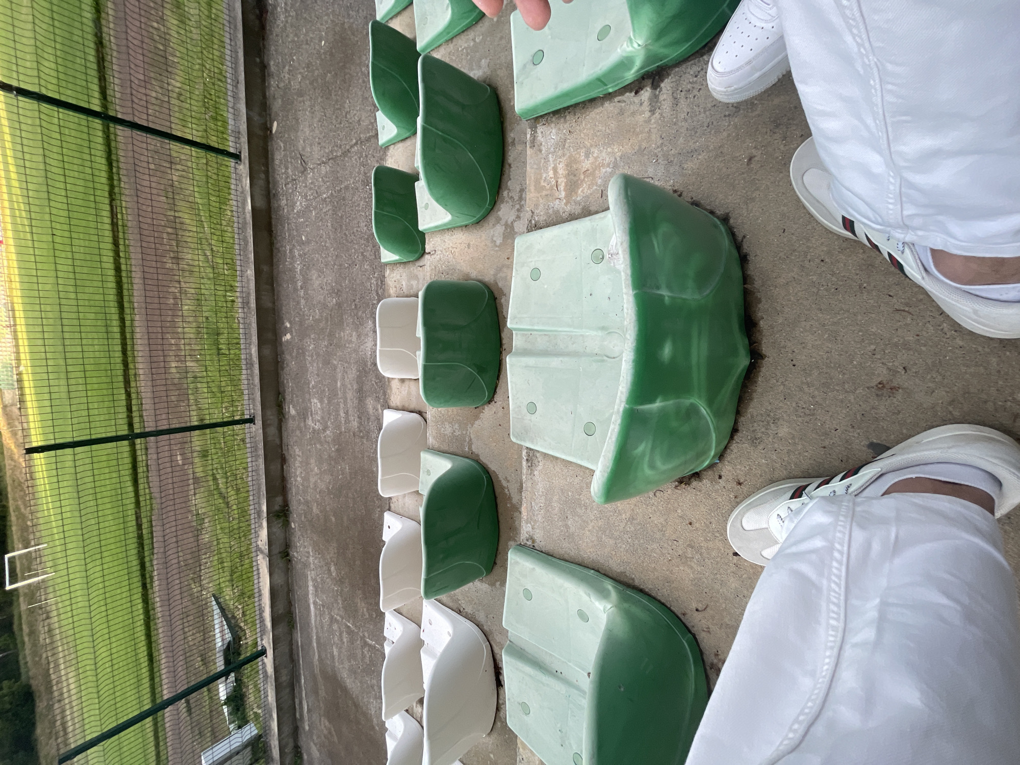 The image captures a ground-level view from a spectator area at an outdoor sports venue, located in Ihtiman, Bulgaria. The foreground is dominated by rows of plastic stadium seats, primarily in shades of green (darker and lighter variations) and interspersed with some white seats. These seats are bolted to a concrete surface that forms the floor of the spectator section.
In the immediate foreground, the lower legs and feet of two individuals are visible. One person wears white trousers and white sneakers with distinctive red and green stripes, accompanied by white socks. Another foot, positioned further up and to the right, wears plain white sneakers, possibly Nike Air Force 1s. A fair-skinned hand with short nails is seen at the top-center, reaching towards one of the green seats.
Beyond the seating area, a dark green wire mesh fence separates the spectators from a large, lush green grassy field, likely a football or sports pitch. White markings are faintly visible on the grass, delineating part of the playing area, possibly a goal box. The lighting on the field suggests late afternoon or early evening, with a soft, warm glow indicating the sun is lower in the sky. The weather appears clear and bright. No discernible text is visible anywhere in the image. The scene suggests a quiet moment at a local stadium or sports complex.
