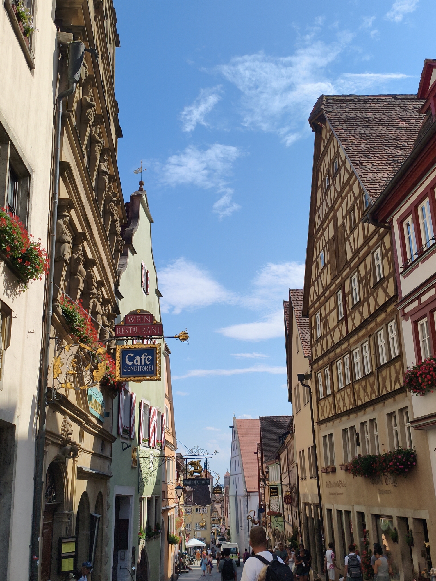 The image depicts a narrow, cobblestone street in Rothenburg ob der Tauber, Germany, lined with historic, half-timbered buildings under a bright blue sky with scattered white clouds. The architecture features intricate wooden beams, red-tiled roofs, and decorative facades, some adorned with statues and flower boxes overflowing with red blossoms.
On the left side of the street, a building displays ornate sculptures above its entrance and a prominent sign that reads "WEIN RESTAURANT" and "Cafe CONDITOREI" in German, indicating a wine restaurant and a cafe/confectionery. Further down the street, other signs for businesses like "Gasthof Greifen" are visible.
Several people are walking along the street, some appearing to be tourists. A man in the foreground is seen from behind, wearing a white t-shirt and a backpack. Other individuals are walking in groups or individually, some interacting with each other. In the distance, more buildings and people are visible, suggesting a lively atmosphere. The scene conveys a sense of old-world charm and the vibrant life of a historic European town.