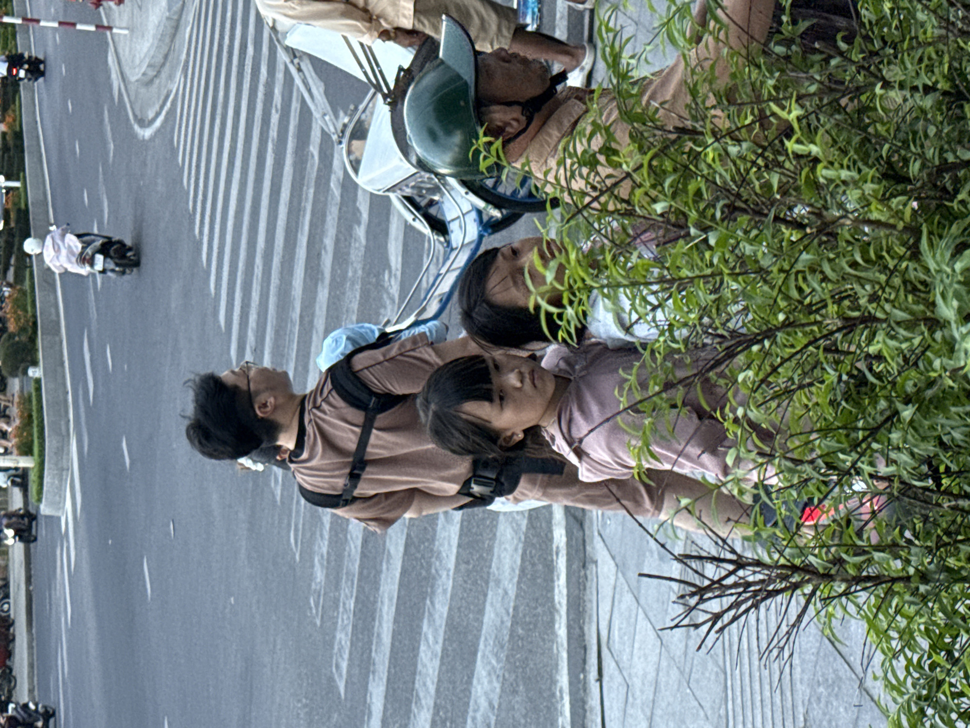 The image captures a street scene in Nha Trang Ward, Vietnam. In the foreground, dense green foliage partially obscures the view. Behind the plants, a man wearing glasses and a light brown t-shirt with a black strap harness stands in profile, facing to the right. He appears to be carrying a child, indicated by a blue cloth visible on his back. A young girl in a pink hooded jacket stands to his left, looking to the side with a neutral expression. To the right of the man, another child with dark hair is partially visible. Behind them, a bicycle-like vehicle with a white canopy is parked on the street. A man wearing a green helmet and a light brown jacket is seated in the foreground of this vehicle. The background features a crosswalk on a paved road with several motorcycles and people.