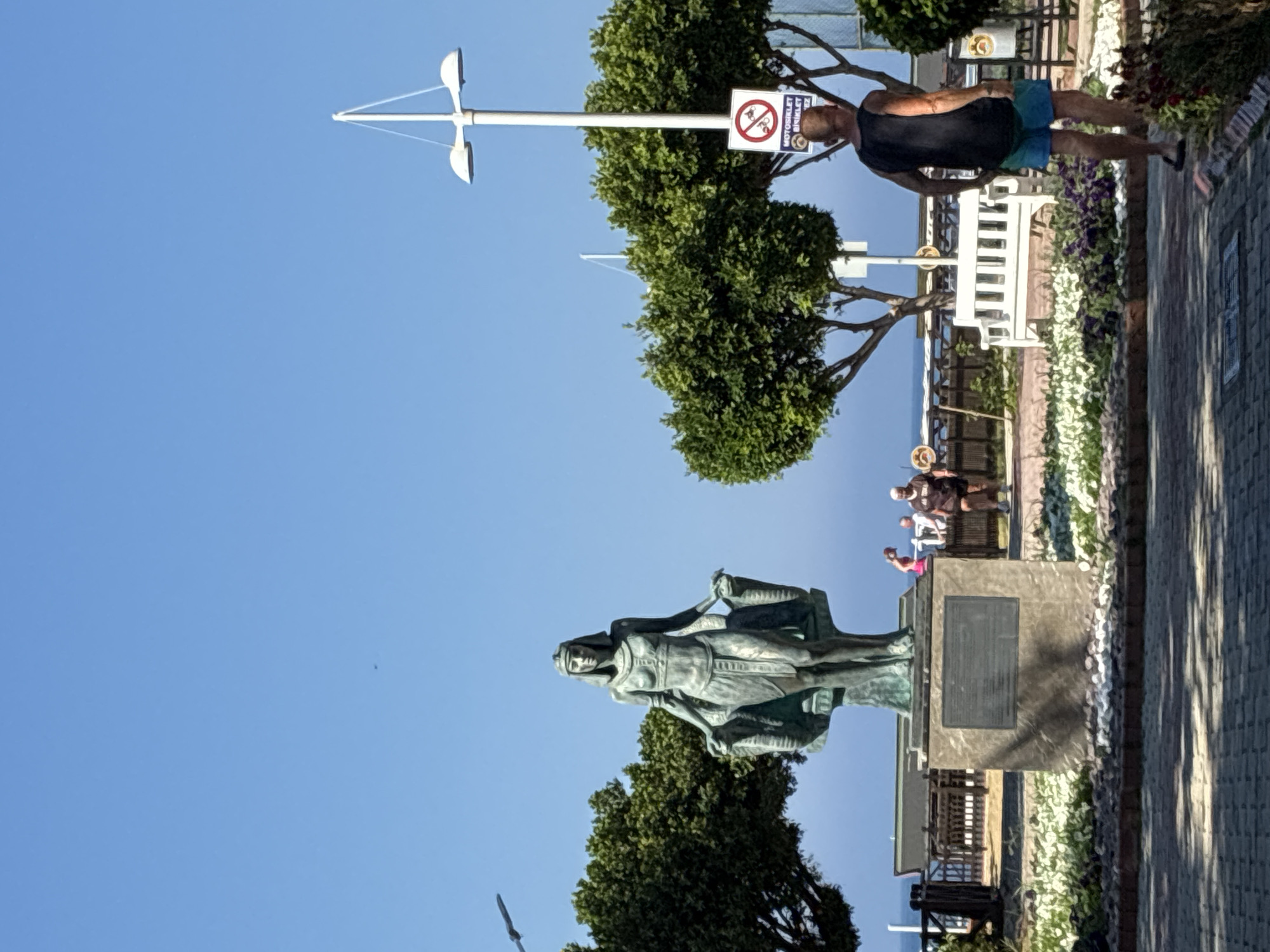 This image captures a daytime scene in Alanya, Turkey, featuring a statue, multiple individuals, and coastal elements under a clear blue sky.
In the central-left foreground, a bronze-colored statue of a female figure, identified as Cleopatra, stands on a rectangular stone pedestal. The figure is adorned with ancient Egyptian-style clothing and a headdress, holding what appear to be two ornate baskets or urns at her hips. A dark rectangular plaque with engraved text is affixed to the front of the pedestal. White and grey pebbles surround the base of the statue, which is itself encircled by white and purple flowering plants within a landscaped bed. The foreground consists of a paved pathway made of interlocking brick pavers.
To the right of the statue, a bald or closely-shaven man, viewed from behind, walks along the paved path. He wears a black sleeveless shirt, blue shorts, and dark flip-flops. Further right, a white, slatted wooden bench is visible amidst green foliage. A tall, white lamppost with two outward-curving light fixtures stands behind the man. Attached to the lamppost is a circular regulatory sign featuring a red border, a diagonal line, and crossed-out icons of a motorcycle and a bicycle, indicating a prohibition. Turkish text below the symbols reads "MOTOSİKLET BİSİKLET İZ".
In the mid-ground, dense green trees frame the scene. Beyond the trees and a wooden fence or railing, the calm, blue sea is visible under a distant horizon. Several additional indistinct individuals are present in the background near the coastline, including one person in a pink top and another in dark clothing. The upper portion of the image is dominated by an uninterrupted, clear blue sky, with a small dark speck, possibly a bird, visible in the upper-left quadrant.