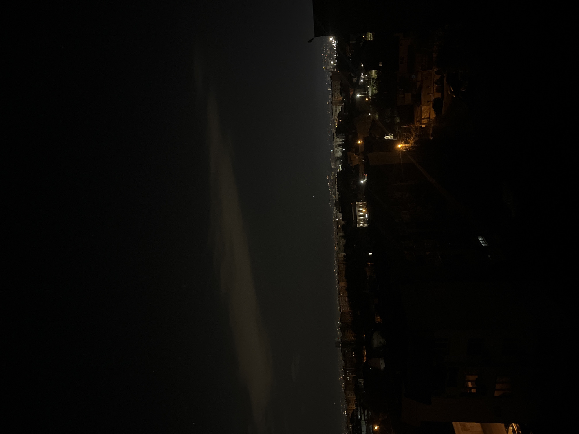 The image is a nighttime, long-range shot of a city skyline. In the lower right corner, there are dark structures suggesting a building or elevated terrain, likely the vantage point of the photographer. Across the center, a body of water or a large expanse of darkness divides the view. The opposite side displays a line of lights, indicating the city's urban development along the horizon. The lights suggest a residential or commercial area with street lighting and lit buildings. The overall environment suggests an urban setting at night, though specific details about the city of Sofia, Bulgaria, are not directly discernible from the image.