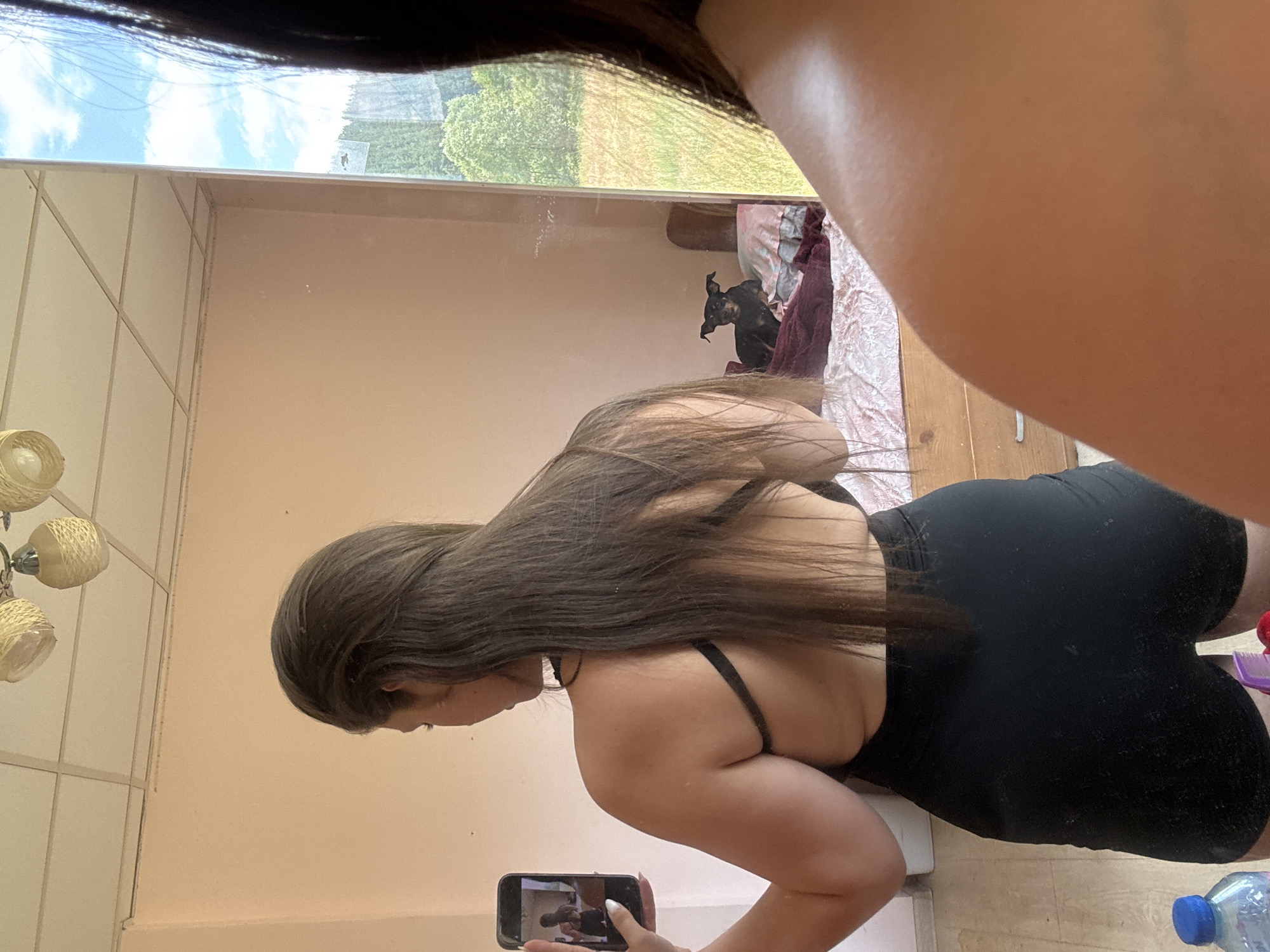 A woman is standing in a room and taking a selfie in a mirror. She is wearing a black sports bra and black shorts. Her long brown hair is down her back. In the background of the mirror reflection, there is a bed with pillows and a dark-colored dog sitting on it. A window is visible in the mirror reflection, showing trees and a sky. The room appears to be a bedroom, with a wooden dresser and a ceiling fan. The floor is wooden, and a blue water bottle is visible on the floor. The image was taken in Prodanovtsi, Bulgaria.