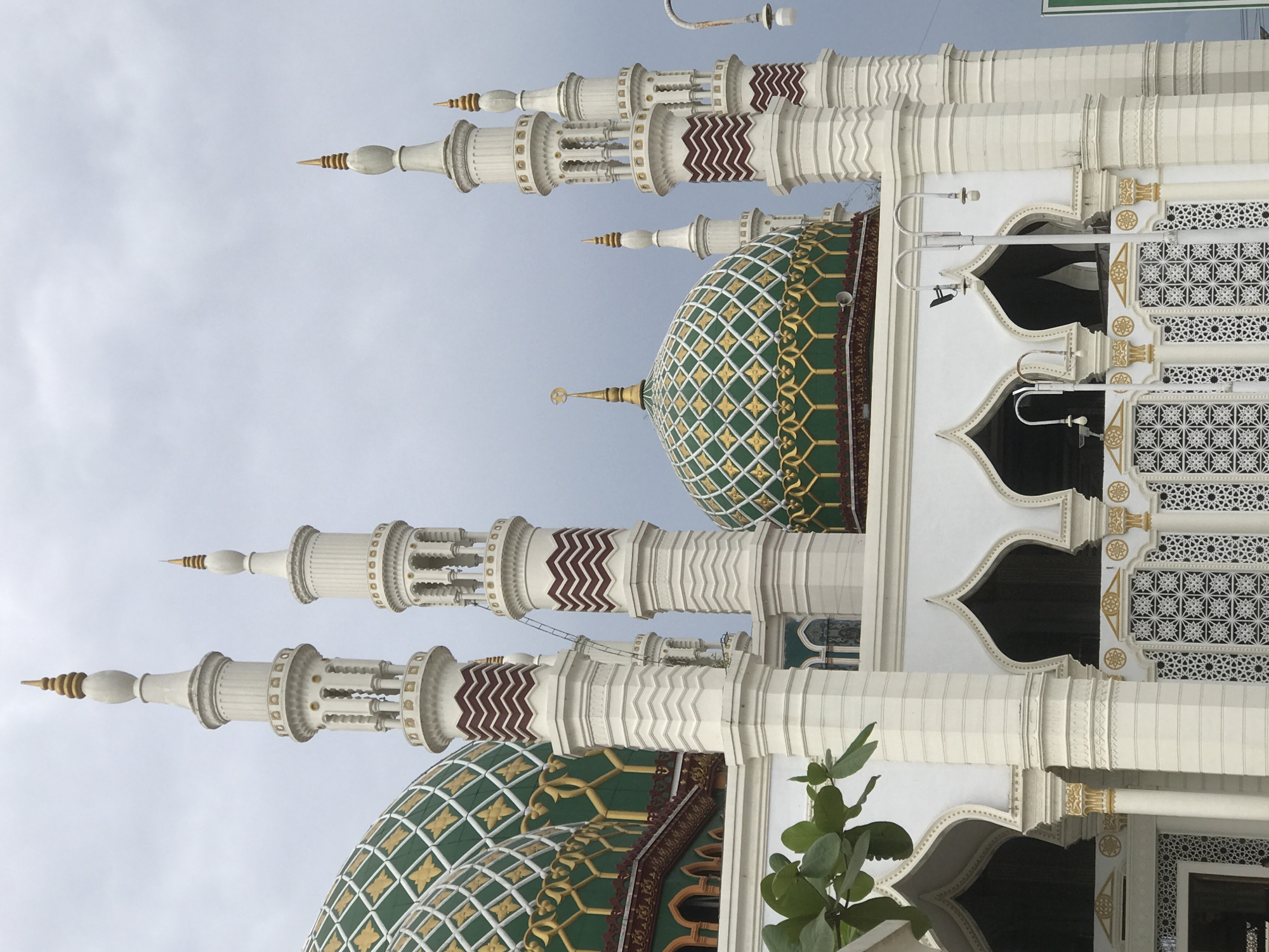 The image shows a mosque with a green and gold dome and white minarets. The sky is overcast. The building is in Kotapantonlabu, Indonesia. The architectural style is ornate. There are plants near the base of the building. The image is rotated.