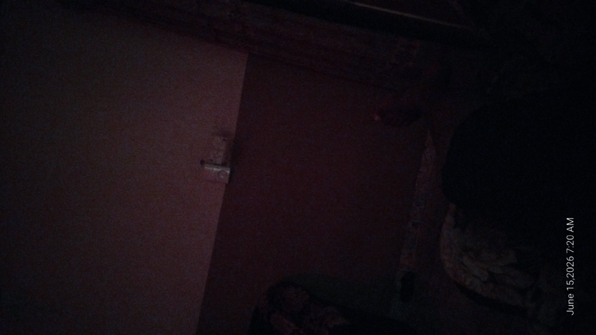 The image is a dimly lit interior scene, likely a bedroom, with a date and time stamp indicating "June 15, 2026 7:20 AM". The overall quality is poor due to low light and noise, making detailed identification of objects challenging. A dark red wall occupies much of the frame. In the upper portion of the wall, a small, white object is mounted, possibly an electrical outlet or switch plate. In the foreground, the lower half of the image is extremely dark, obscuring most details, but hints of bedding or fabric with a pattern are visible on the left. The location in Howayi, Nigeria, is not discernible from the visual information in the image.