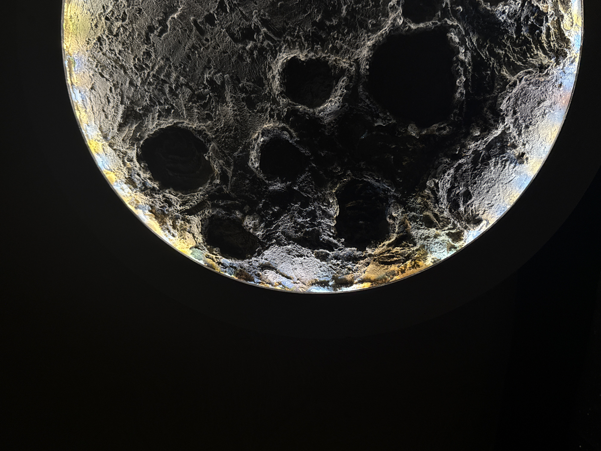 The image depicts a lunar-like sphere with a cratered surface illuminated from behind, creating a stark contrast against a dark background. The sphere is likely an art installation or model of the moon. The location in the city of Chaikovsky, Russia, is not evident from the image alone.