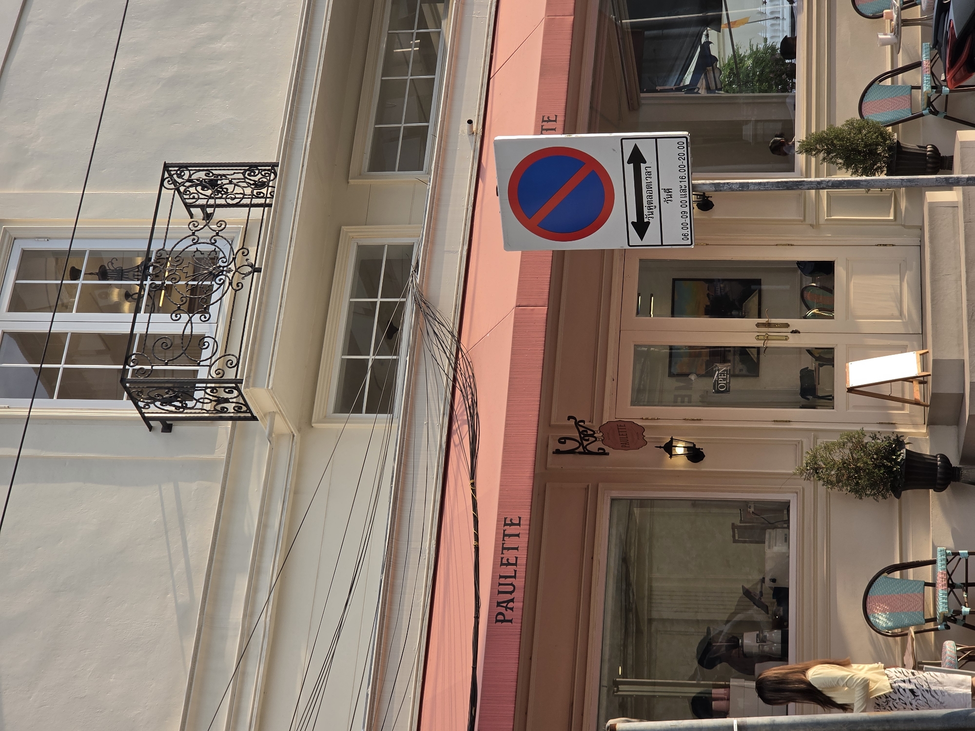 The image depicts a street scene in Bangkok, Thailand. The main focus is the storefront of a business named "PAULETTE," indicated by the awning and signage. A traffic sign indicating "no parking" during specified hours in Thai and English is present. The building features architectural details, including a small balcony with ironwork. Outside the storefront, there are potted plants and chairs, suggesting an outdoor seating area. A person is visible in the lower part of the image. Power lines are visible against the building's facade. The building's facade is a mix of pale yellow and pink. The photo is taken during daylight hours.