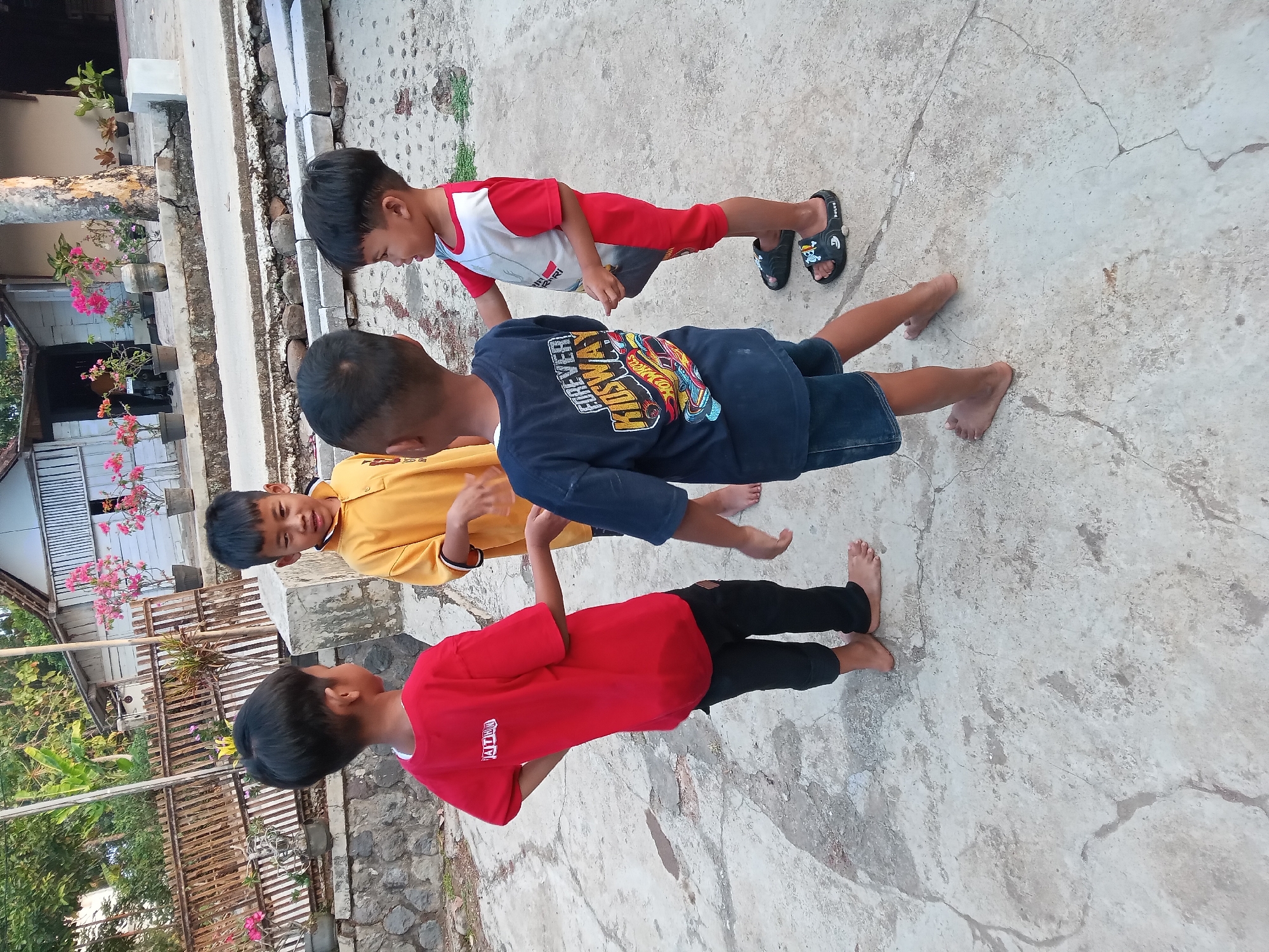 Four young boys are standing in a circle on a paved outdoor area. The boy on the left is wearing a red t-shirt and black pants. The boy next to him, facing him, is wearing a yellow polo shirt. The boy in the center is wearing a dark blue t-shirt with a graphic print and denim shorts. The boy on the right is wearing a red and white t-shirt with red shorts and black sandals. They appear to be interacting with each other, possibly playing a game. In the background, there is a rustic wooden fence, a building with pink flowers in pots, and trees. The ground is made of cracked concrete. The scene suggests a casual outdoor gathering in a residential area. This appears to be in Kedunggudel, Indonesia, as indicated by the surrounding architecture and vegetation.