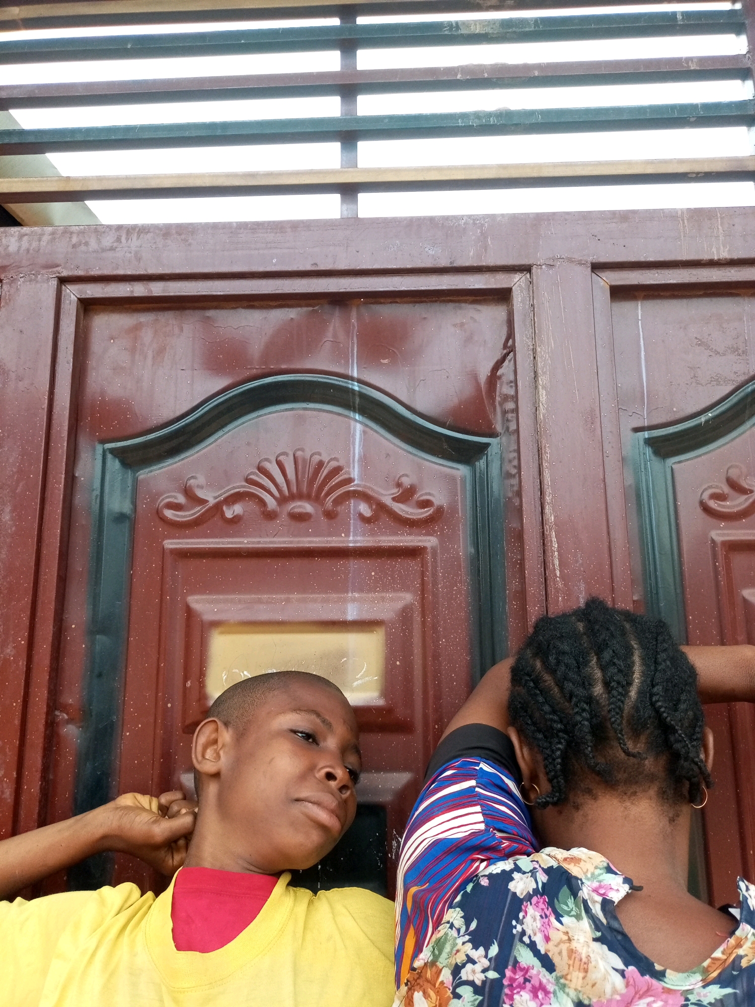 In Tempure, Nigeria, two young individuals stand against a dark brown wooden door adorned with black trim and decorative elements. The individual on the left, facing away from the camera, has short hair and wears a yellow t-shirt with a red underlayer. Their left arm is raised behind their head, touching their neck. The individual on the right, also facing away, has braided dark hair and wears a floral-patterned top and a striped sleeve. Both individuals appear to be near a slatted metal structure above the door. The lighting suggests it is daytime.