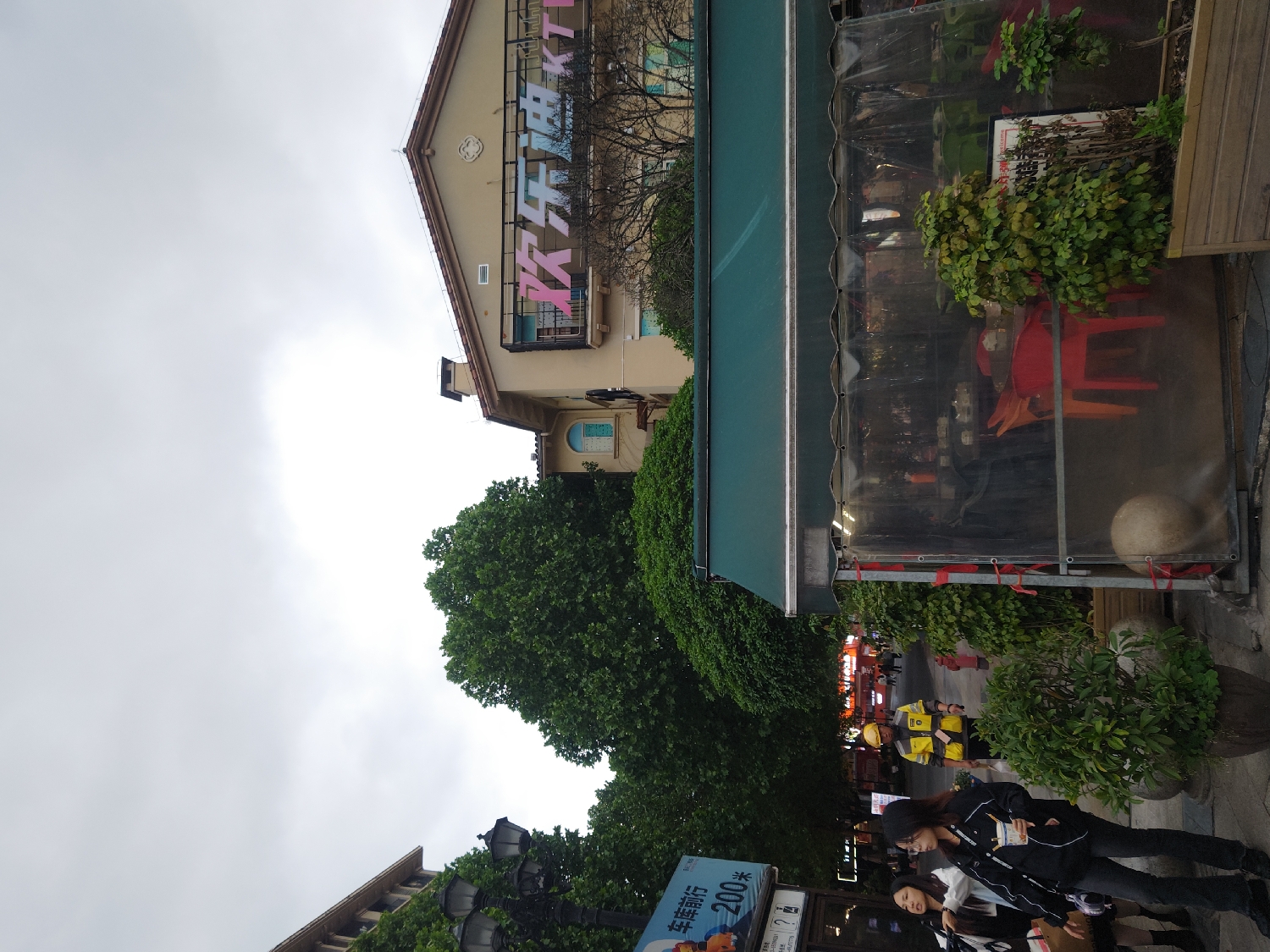This image depicts an urban street scene in Chongqing High-tech Zone, China, under an overcast sky. The light grey sky suggests cloudy weather during the daytime.
In the background, a light-colored building with a sloped roof features large, colorful Chinese characters prominently displayed on its upper floor. The characters "欢笑" (huānxiào), meaning "laughter" or "joyful," are visible in pink and blue. A large, dense green tree stands in front of this building, partially obscuring its lower sections.
In the mid-ground, an outdoor dining or seating area is covered by a green awning and enclosed by transparent plastic sheeting, secured with red ties. Inside, red and orange chairs are arranged, suggesting a café or restaurant. Several potted green plants are placed around this outdoor section and along the sidewalk.
On the paved sidewalk in the foreground, two young women are walking or standing. One woman has long dark hair and wears a dark jacket, dark pants, and a beanie, holding an item in her hand. The other, next to her, wears a white top and a dark outer garment. Further down the street, several individuals, some appearing to be delivery riders in yellow uniforms, are visible on what look like scooters or bikes.
A blue and white street sign is present on the left, displaying Chinese text: "车库前行 200米" (Chēkù qiánxíng 200 meters), which translates to "Garage straight ahead 200 meters." A street lamp post is also visible. The scene portrays a bustling commercial area with pedestrian activity and urban infrastructure.