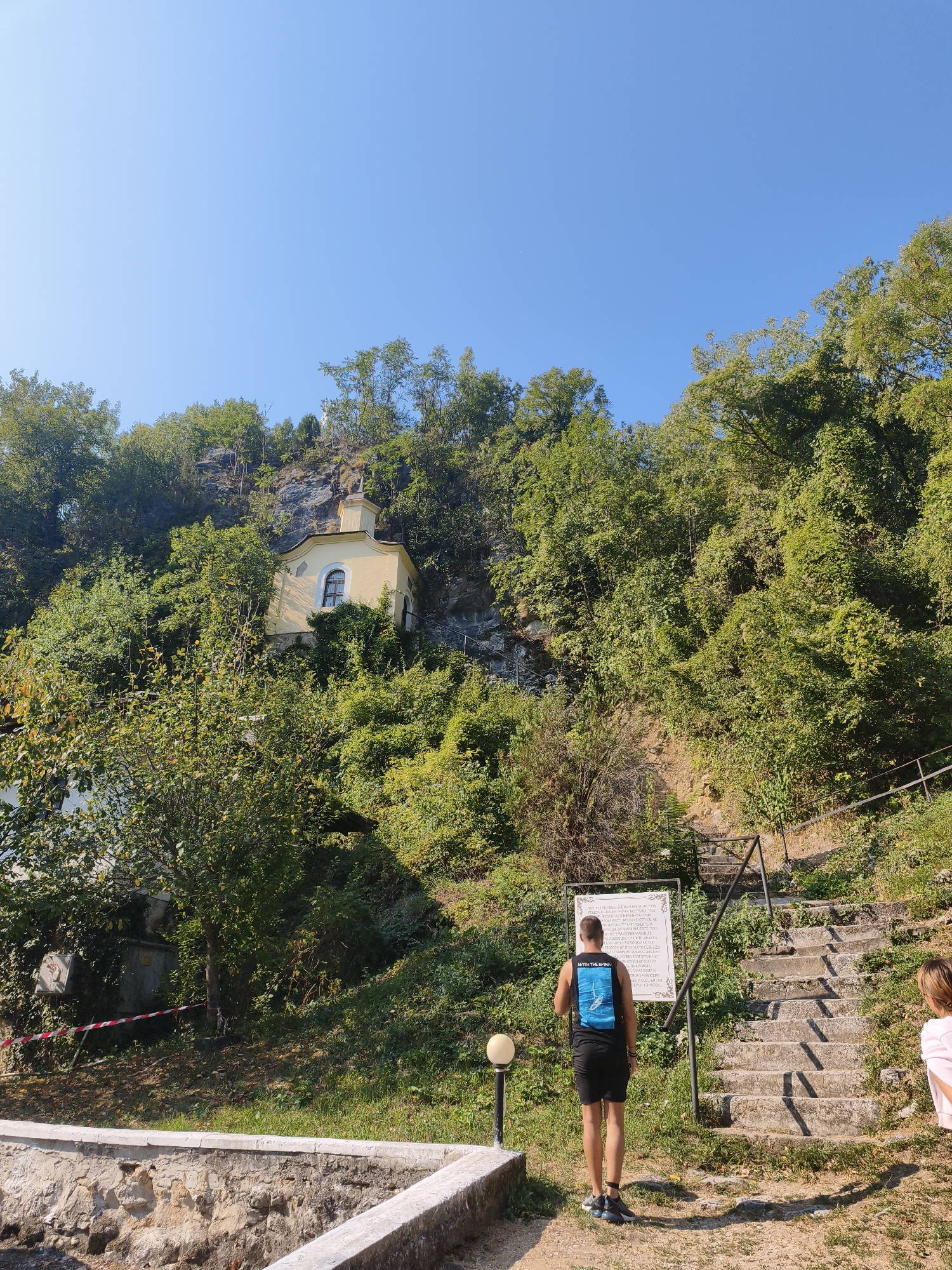 The image depicts the Vratsata Gorge, near Lyutibrod, Bulgaria. A small, yellow chapel is built into the side of a rocky cliff, surrounded by lush green trees. In the foreground, a set of stone stairs leads up the hillside, with a railing for support. A man wearing a black tank top and shorts stands at the bottom of the stairs, facing away from the camera, looking at a white informational sign. A young girl in a pink top is visible to the right of the stairs, partially obscured by the railing. The sky is clear and blue.