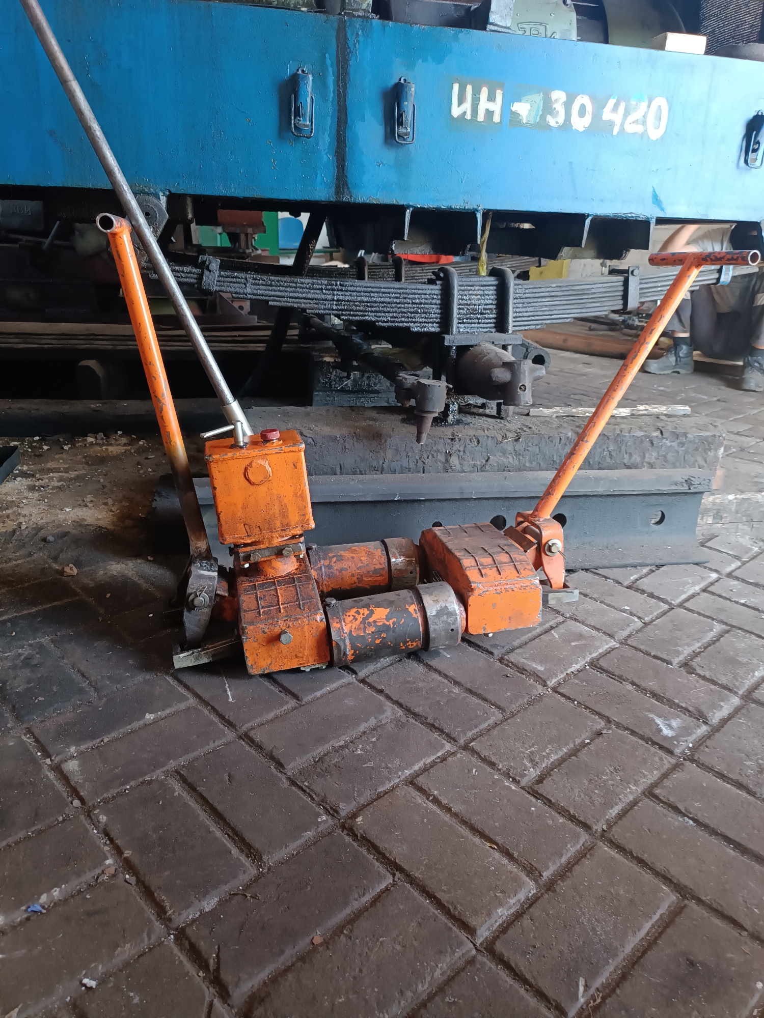 In Vuhlehirsk, Ukraine, an industrial setting is depicted where two heavy-duty, orange hydraulic jacks are positioned beneath a large blue mechanical component, possibly part of a train or industrial machinery. The jacks are engaged, indicating they are actively supporting or lifting the weight. One jack has its operating lever extended upwards, while the other's lever is in a lower position. The machinery above shows a visible identification marking: "UH 30 420" in white lettering. The ground is paved with dark, interlocking bricks, and dust and debris are present, suggesting an active work area. In the background, partially visible, are what appear to be the legs of at least one person wearing dark trousers and boots, indicating human presence in the vicinity, likely involved in maintenance or operation. The overall scene is one of industrial activity, focused on heavy machinery maintenance.