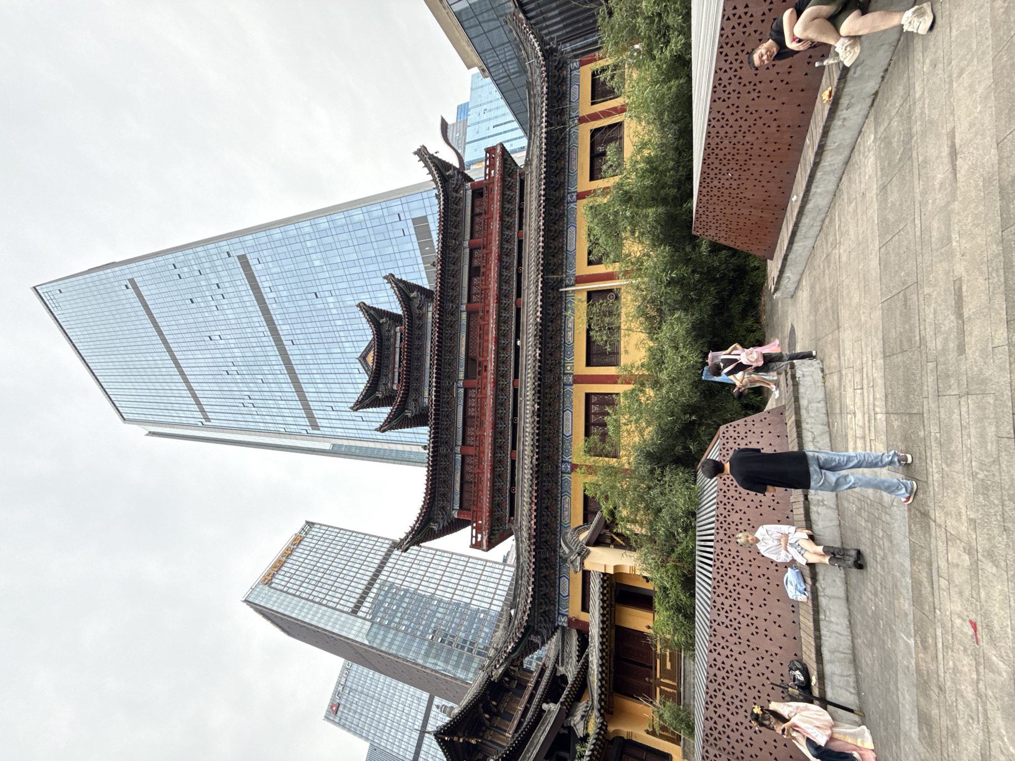 The image shows a juxtaposition of traditional Chinese architecture with modern skyscrapers in the Yuzhong District of Chongqing, China. A multi-tiered, elaborately decorated traditional building stands prominently in the center of the frame, with its dark, ornate eaves and colorful detailing. Behind it, a towering glass-clad skyscraper rises, reflecting the sky, creating a stark contrast between old and new. Other skyscrapers can be seen in the background.
On the right side of the image, there is a textured wall with triangular cutouts, alongside a pedestrian area. Several people are present: one is lying down, others are standing, walking, or seated. The ground is paved with stone. The overall environment is urban and appears to be a public space where modern life intersects with historical preservation.