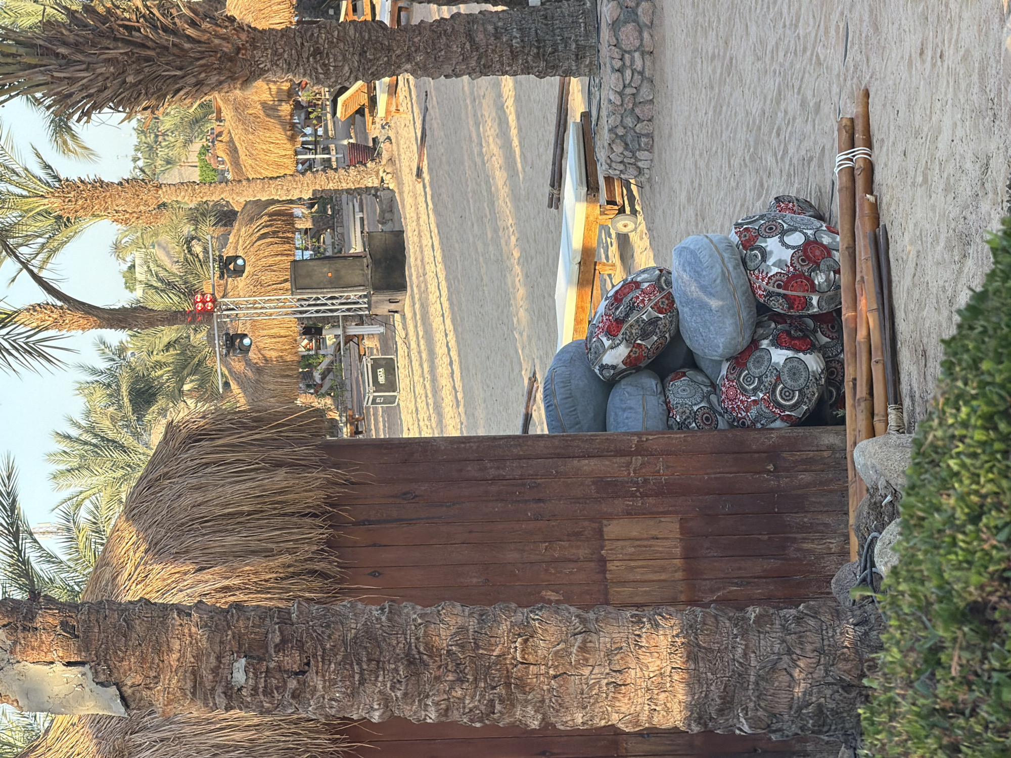 The image depicts a tropical beach scene in Sharm El Sheikh, Egypt, likely at a resort. In the foreground, a large palm tree trunk is prominent, leaning against a wooden structure with a thatched roof. Next to this structure, a stack of large, round cushions is piled up on the sandy ground. Some of the cushions have a red and white patterned design, while others are solid blue-gray. Bundles of bamboo poles are also present on the sand, tied with white rope.
In the mid-ground, there are more thatched-roof structures, possibly cabanas or bar areas, and several lounge chairs. A sound system, including a large speaker and a lighting rig, is set up on a metal frame, suggesting preparations for an event or entertainment.
The background reveals more palm trees, sandy terrain, and glimpses of buildings and possibly hills in the distance. The overall lighting suggests it is either early morning or late afternoon, with warm sunlight casting shadows on the sand. There are no people visible in the image. The environment is sandy and dry, characteristic of a desert coastal area like Sharm El Sheikh.