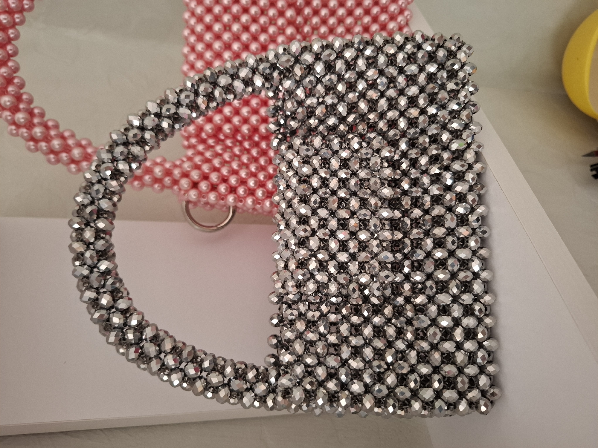 The image displays two beaded handbags on a white shelf. The foreground handbag is silver and is made of faceted beads, creating a sparkling effect. It has a rounded handle also covered in silver beads. The background handbag is pink and appears to be made of smaller, rounder beads. A metal ring is visible attached to the handle of the silver handbag. The lighting suggests an indoor setting. The location of this scene in Korenovsk, Russia cannot be determined from the image.