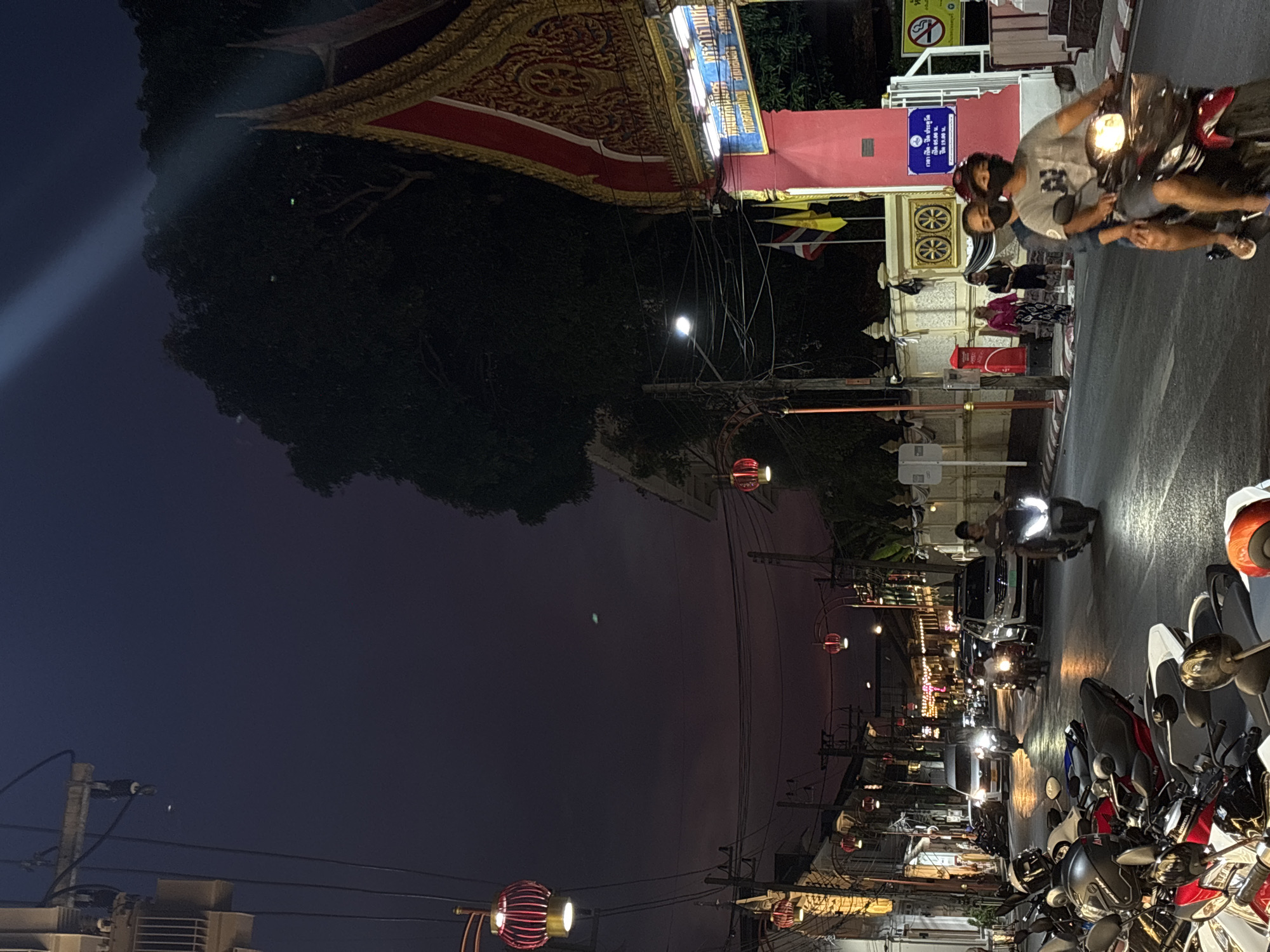 The image shows a street scene in Phuket City Municipality, Thailand, at night. A Thai-style building structure with intricate details is visible on the left side of the street, and a sign is displayed on the building's facade. The sign features Thai script and English text, with opening and closing hours mentioned.
Several motorbikes are parked along the right side of the road. A scooter with two people riding it is seen on the road near the building. There are people standing and walking on the side of the road. Streetlights and hanging lanterns illuminate the street. The sky is dark, suggesting that it is nighttime.