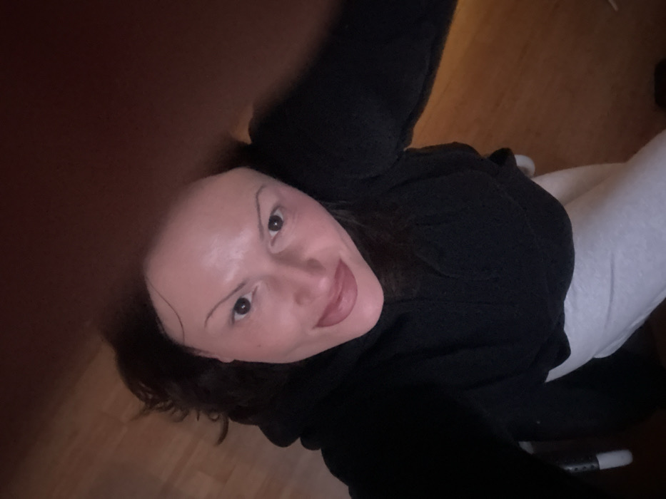 A selfie from a low angle shows a person with fair skin, dark hair, and arched eyebrows. They wear a dark hooded top and light-colored bottoms. The background features a wooden floor. A portion of the image is obscured by a blurry object, possibly a finger. The person is smiling slightly.