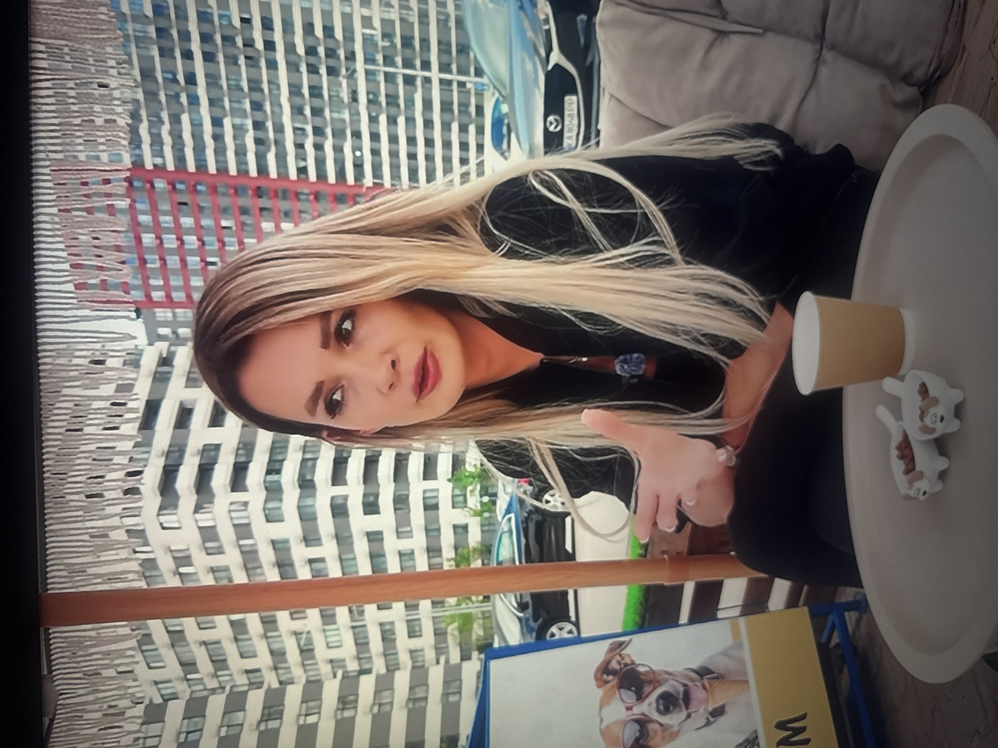 The image shows a woman with long blonde hair sitting in an outdoor seating area. She is wearing a black jacket and looking to her left. In front of her is a round table with a paper cup and two small dog-shaped dishes containing food. To her left, there is a sign with a picture of a dog wearing sunglasses and an ice cream cone. Behind her, there are modern buildings and parked cars. The woman is gesturing with her right hand. The scene appears to be a cafe or restaurant with an urban background. The context suggests this location is in the city of Herzliya, Israel, possibly at a dog-friendly establishment given the dog imagery and food dishes.