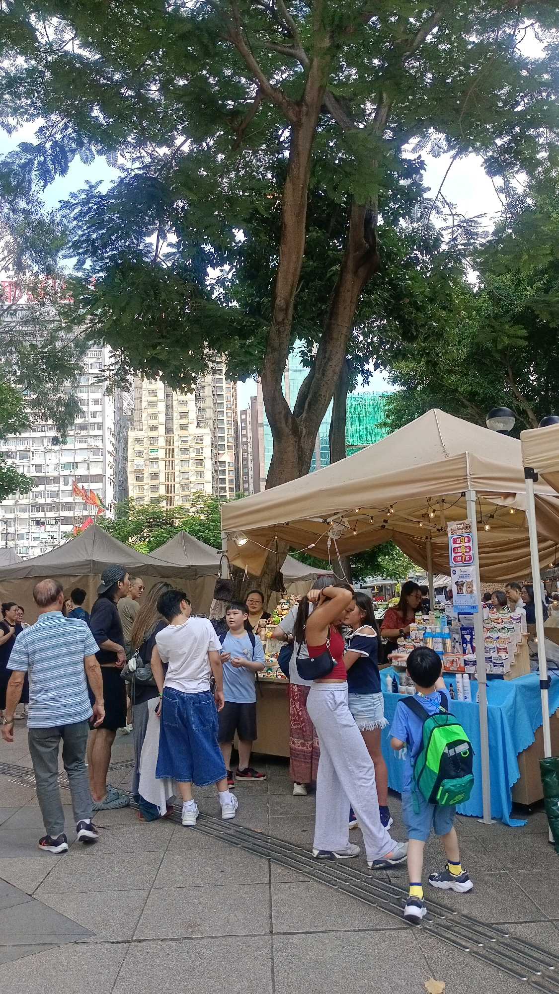 This image depicts an outdoor market scene, likely in a public space in Hong Kong, China, given the architecture of the surrounding buildings. In the foreground, several people are browsing or walking through the market. To the left, a man in a striped shirt and grey pants walks away from the camera. To his right, a group of individuals, including a person in a black cap and dark clothing, and another in a white t-shirt, are visible.
The market itself consists of several stalls with beige canopies. One stall in the center is well-lit with string lights and displays various items on tables covered with a blue cloth. A woman in a red top and grey pants is standing at this stall, facing away from the camera, and a young boy in a blue shirt and backpack is looking at the items.
Behind the market stalls, tall residential buildings with numerous windows are visible, characteristic of urban Hong Kong. Trees with dense foliage provide shade over the market area. The ground is paved with stone tiles. The overall impression is of a lively, casual gathering in a bustling urban environment.