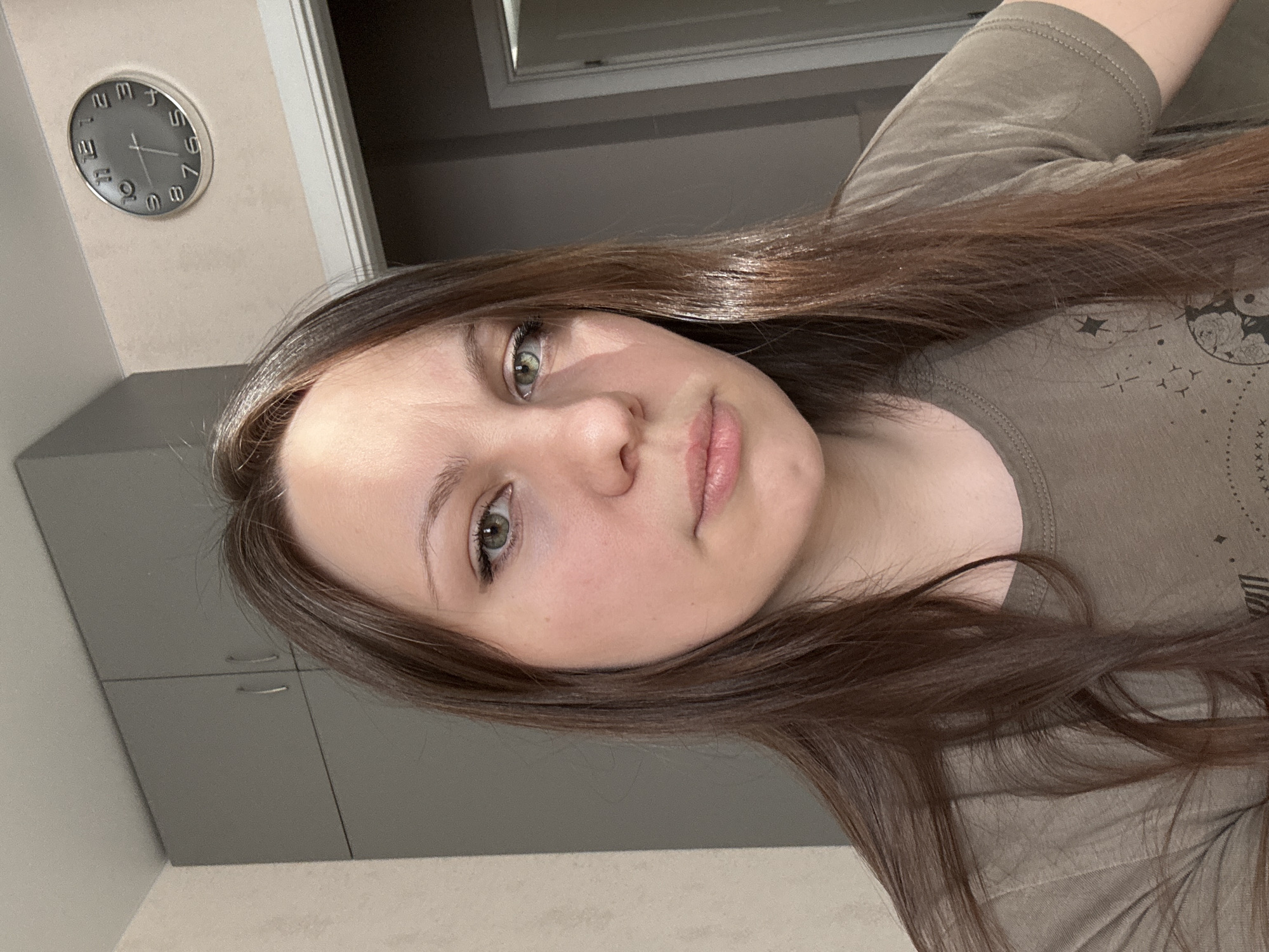 The image shows a woman with long brown hair and green eyes, looking directly at the camera. She is wearing a brown t-shirt. The background is a domestic interior with a grey cabinet on the left and a wall with a clock on the right. A doorway is visible on the far right. There is no indication of the location being Saint Petersburg, Russia.