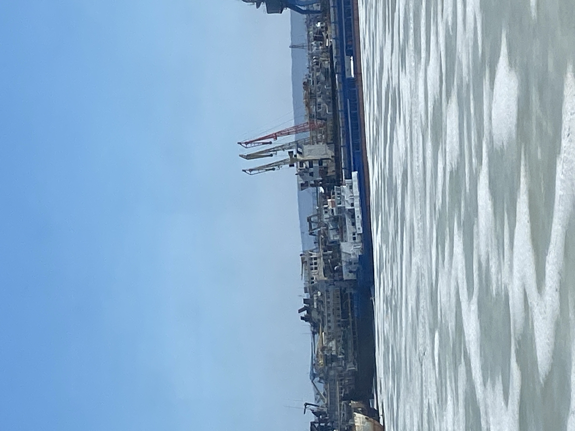 The image shows a view of the Volga River in Chaikovsky, Russia, with a shipyard visible along the shoreline. Several large cranes are present, indicating industrial activity related to shipbuilding or maintenance. The water of the Volga displays a textured surface, reflecting light. The sky is clear and blue.