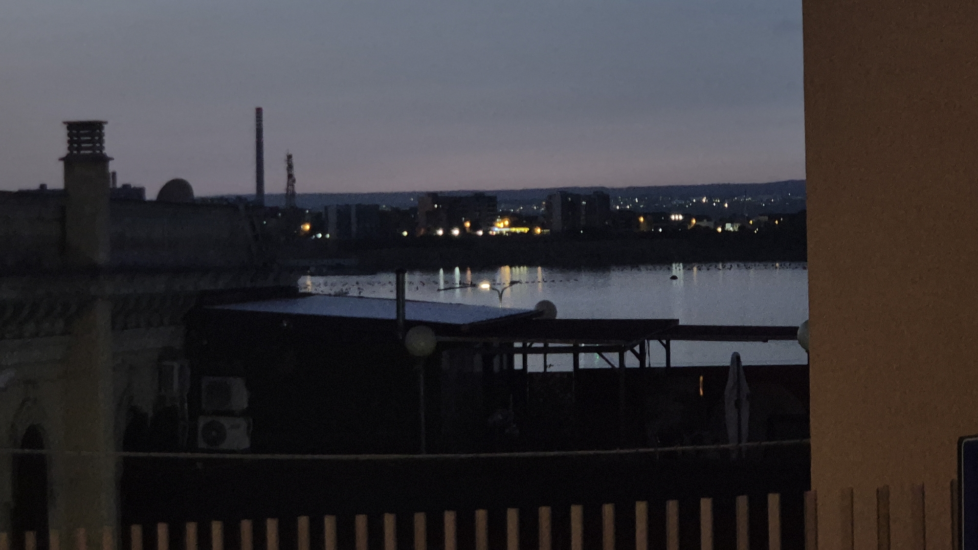 The image depicts a twilight scene overlooking a body of water, likely the Mar Piccolo, in Taranto, Italy. In the foreground, a partially visible structure, possibly a building or rooftop, is on the right, with a light brown, textured surface. To the left, a fence with vertical wooden slats is visible, extending across the bottom of the frame. Behind the fence, a low, dark structure with a corrugated roof is partially obscured by the shadows. Two outdoor air conditioning units are mounted on a dark wall. Further back, the calm surface of the water reflects the faint lights of the city in the distance, creating a blurred glow. The distant cityscape is characterized by a collection of buildings and industrial structures, including a prominent smokestack and a domed building. The sky is a deep, muted blue, indicative of dusk or dawn. A single figure, appearing to be a person wearing a draped garment, stands in the middle ground, adding a mysterious element to the otherwise still landscape.