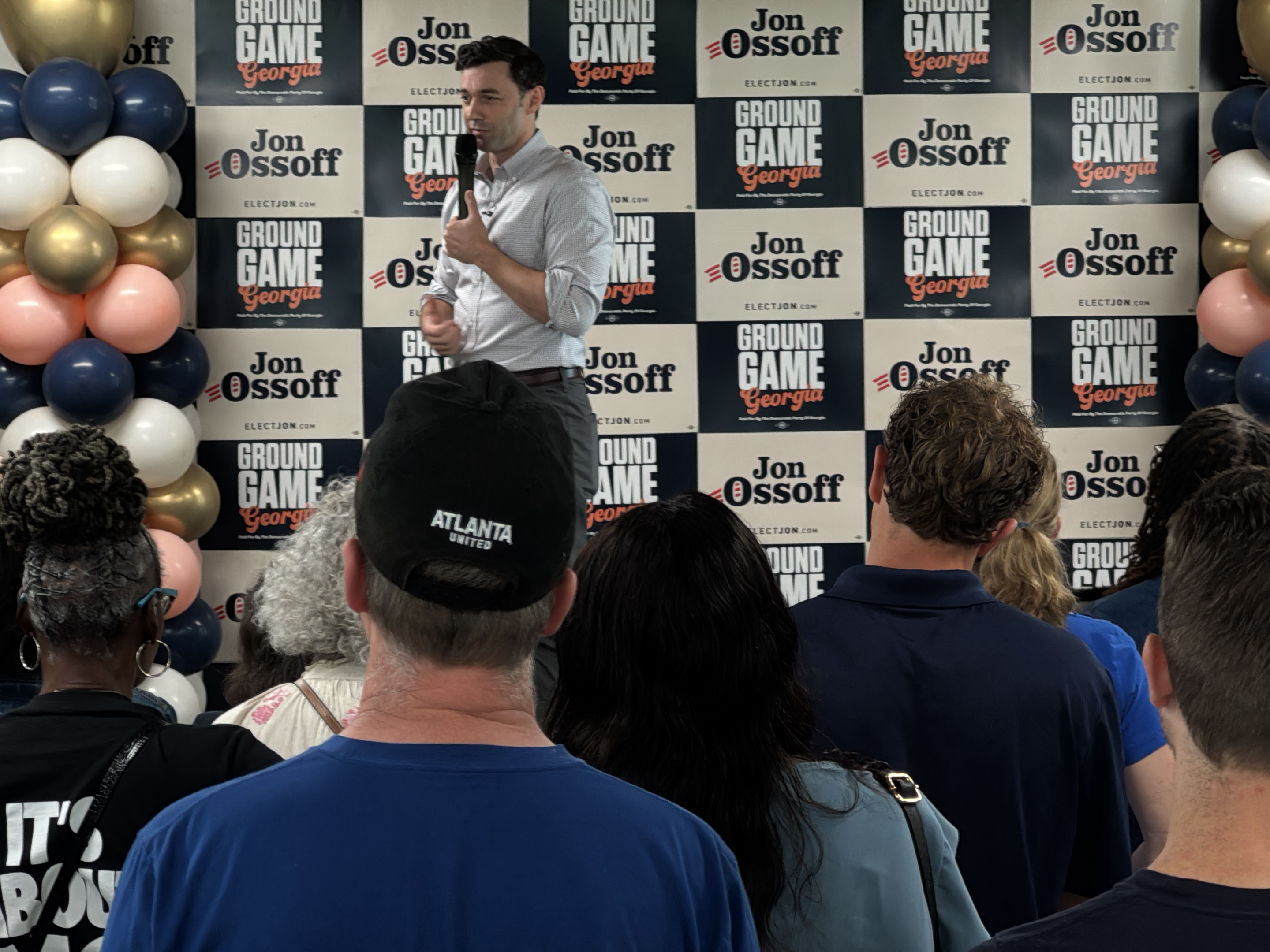 The image shows Jon Ossoff, an American politician, speaking to a crowd of people. He is standing behind a podium with the words "GROUND GAME Georgia" and "Jon Ossoff ELECTJON.COM" printed on it in a repeating checkered pattern. Balloons are arrayed on either side of the backdrop. The crowd is diverse, with people of various ages and ethnicities. An "Atlanta United" hat is clearly visible. The setting appears to be an indoor event space. The location is Decatur, Georgia, United States, based on the campaign signage visible.
