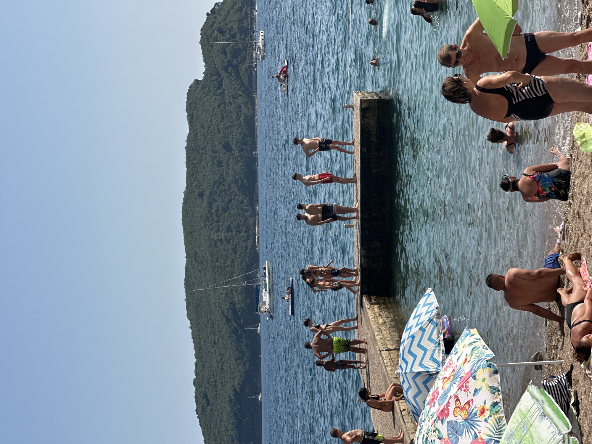 The image captures a sunny day at a beach in Budva, Montenegro, with a pier extending into the calm blue sea. The pier is populated by a group of people, some standing and observing the water, while others are engaged in conversation or simply relaxing. Children are also present, some standing near the edge of the pier. In the background, a lush green, tree-covered island rises from the sea, with several sailboats and a larger white boat cruising on the water. On the shore in the foreground, beach umbrellas provide shade to beachgoers. One person is lying on a towel on the sand, sunbathing, while another is seated, facing the sea. Several other individuals are visible in the shallow water. The overall scene depicts a lively and relaxed atmosphere characteristic of a summer day at a coastal resort.
