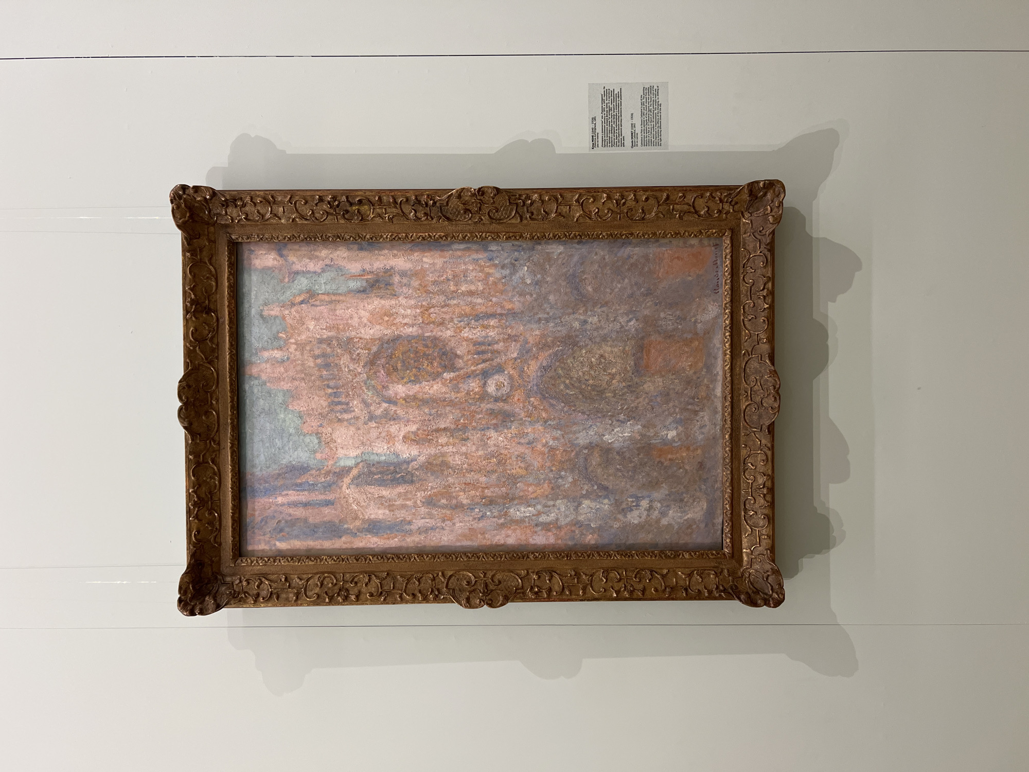 A framed painting hangs on a white wall. The painting appears to be an impressionistic landscape with dominant colors of pink, brown, and muted blues. The frame is ornate, golden-brown with intricate detailing. A small label is visible above the painting.
The location in the city of Belgrade, Serbia is not determinable from the image.