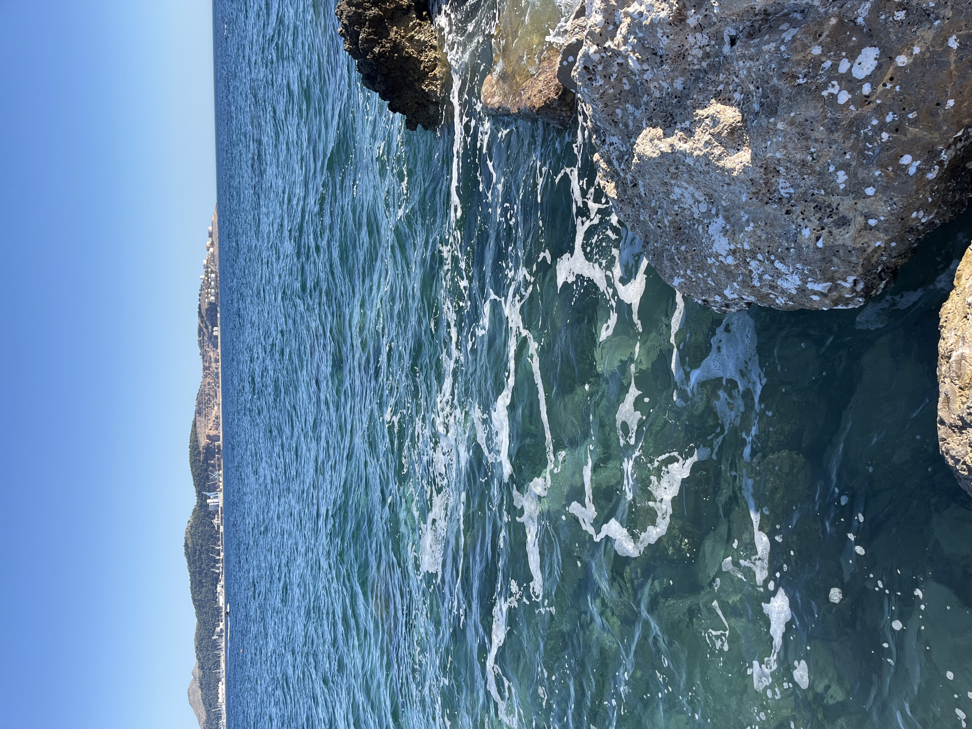The image depicts a clear blue sky over the Adriatic Sea in Bar, Montenegro. The foreground features rocky shoreline with large, weathered boulders. The water is turquoise and transparent, revealing the rocky seabed beneath. White foam from the gentle waves creates intricate patterns on the water's surface. In the distance, a peninsula with lush green hills and a coastline dotted with buildings and industrial structures is visible. A small boat can be seen navigating the sea. The scene conveys a serene coastal environment with a hint of industrial presence on the horizon.